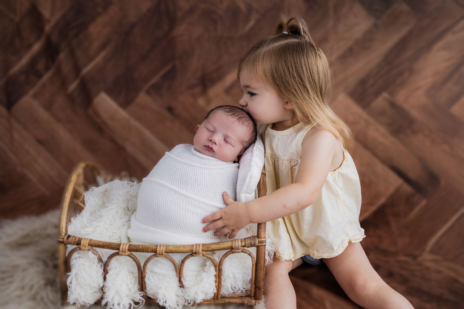 Young child lovingly embraces swaddled newborn baby sibling resting in rustic wicker basket against brown backdrop.