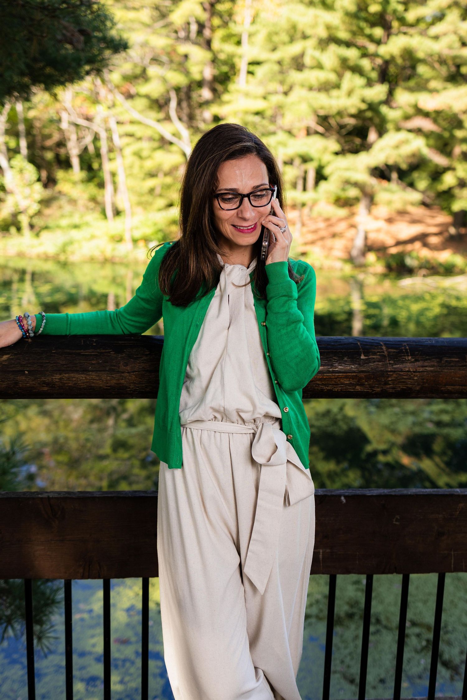 Person wearing green blazer and white dress leans on wooden railing in sunny outdoor setting.