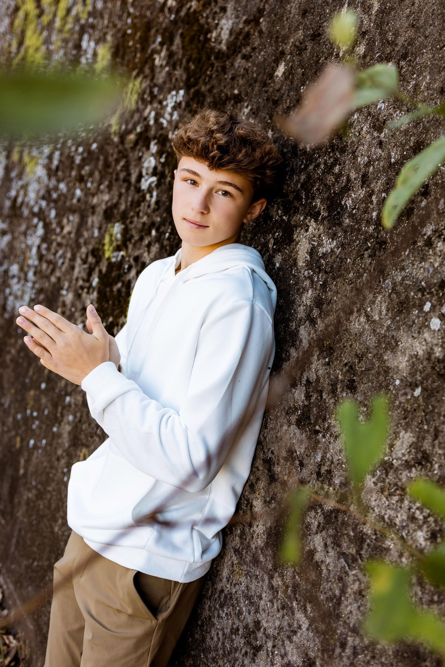 A relaxed outdoor pose against a mossy tree trunk wearing a white sweatshirt.