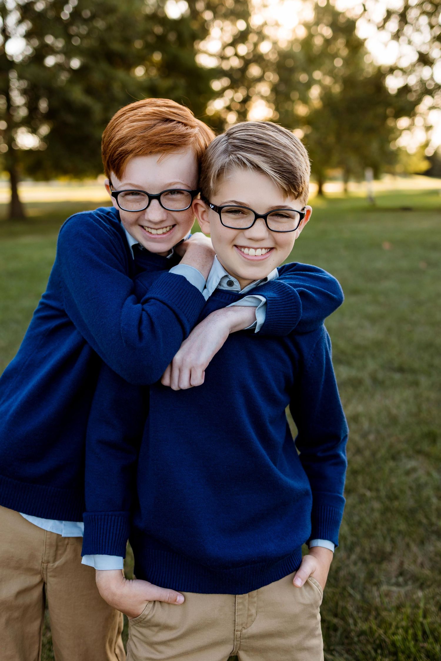Two children in navy blue sweaters and khaki pants share a playful hug while smiling outdoors at sunset.