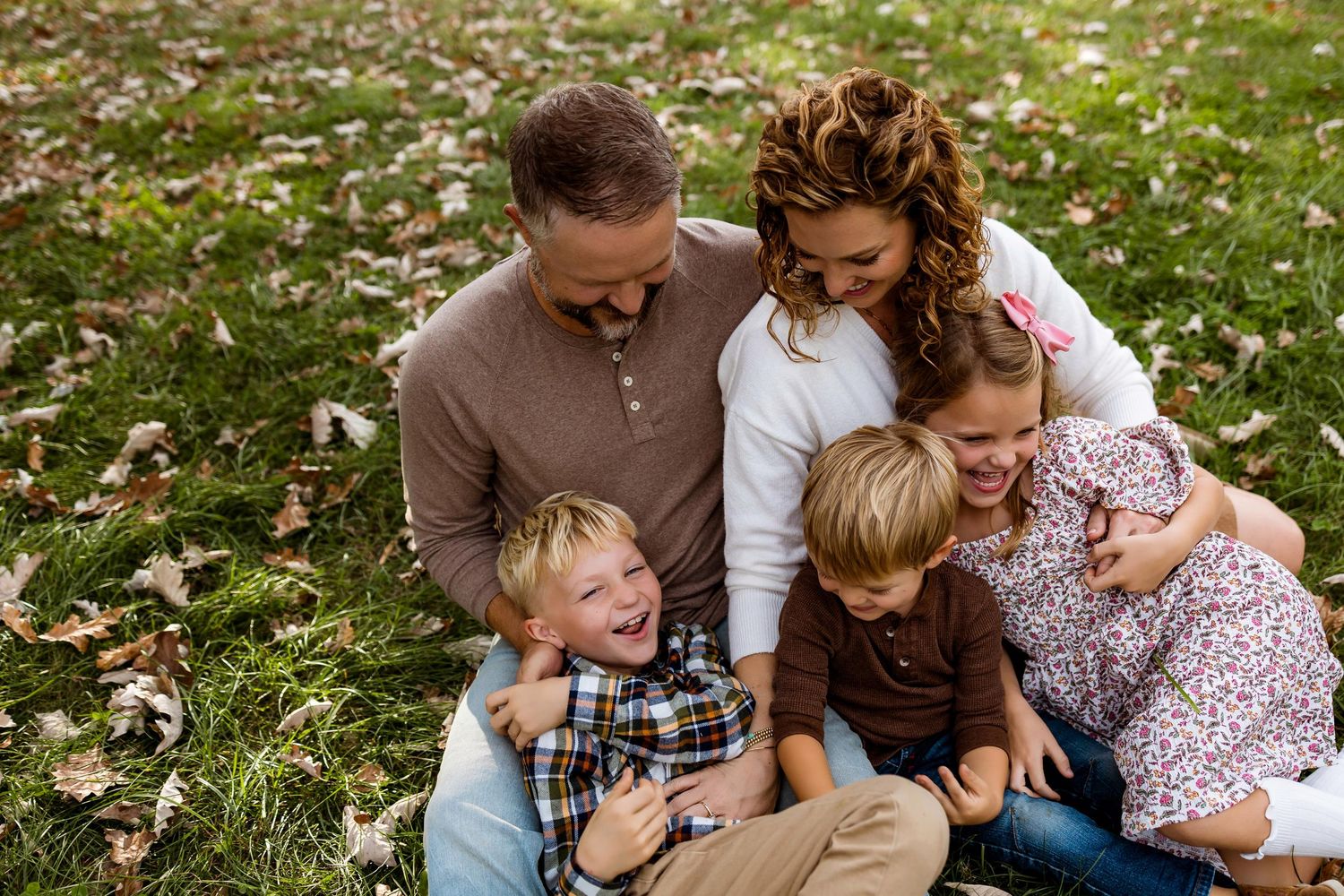 Family sits together on grass covered in fallen autumn leaves during an outdoor photo session.