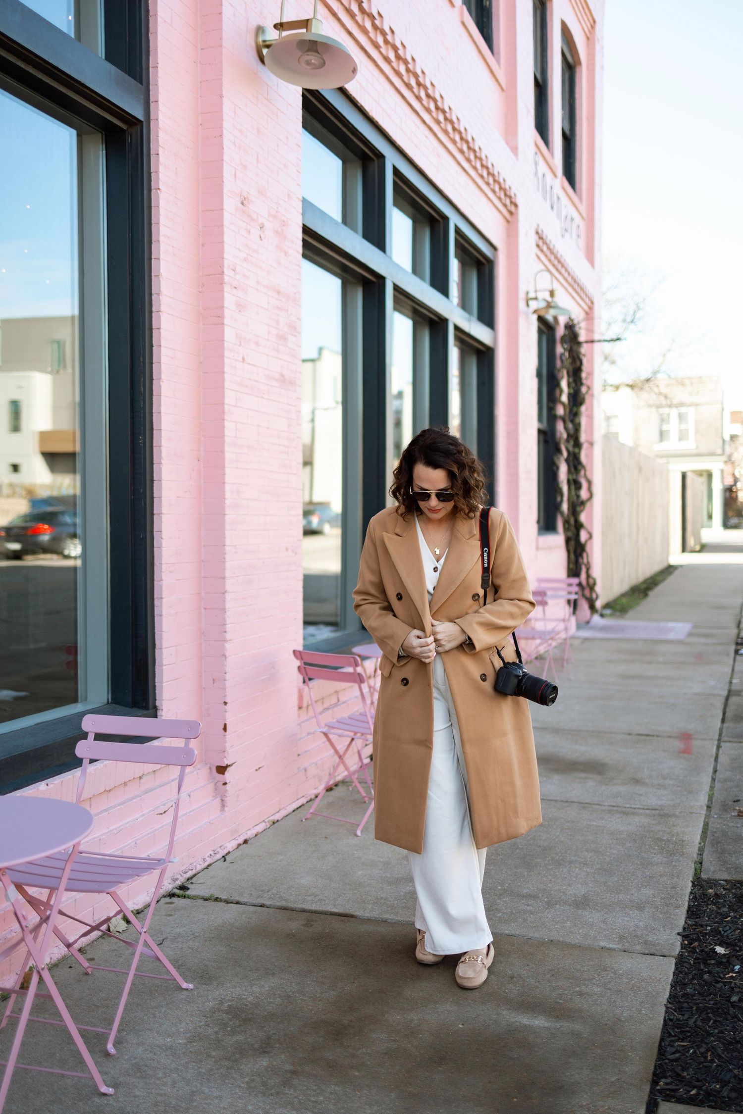 Stylish camel coat and white pants outfit styled against a pink building facade with pink bistro chairs.