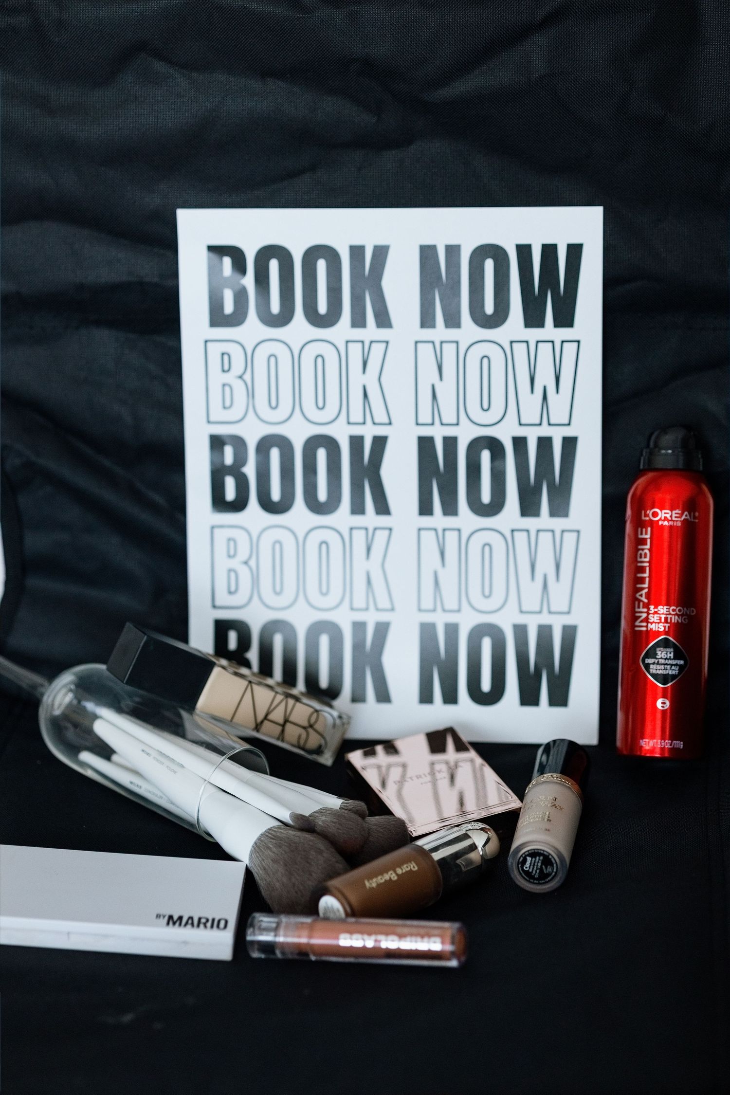 Makeup products including NARS, L'Oreal, and Mario Badescu arranged beside a bold black and white Book Now sign.
