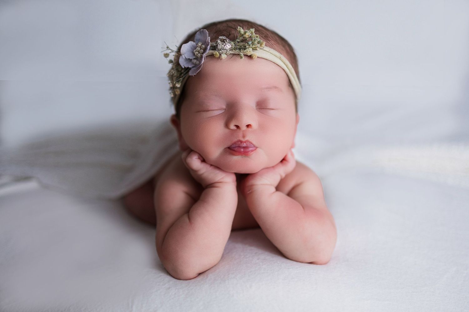 Sleeping newborn baby with delicate headband lying on white blanket with hands under chin in studio portrait.