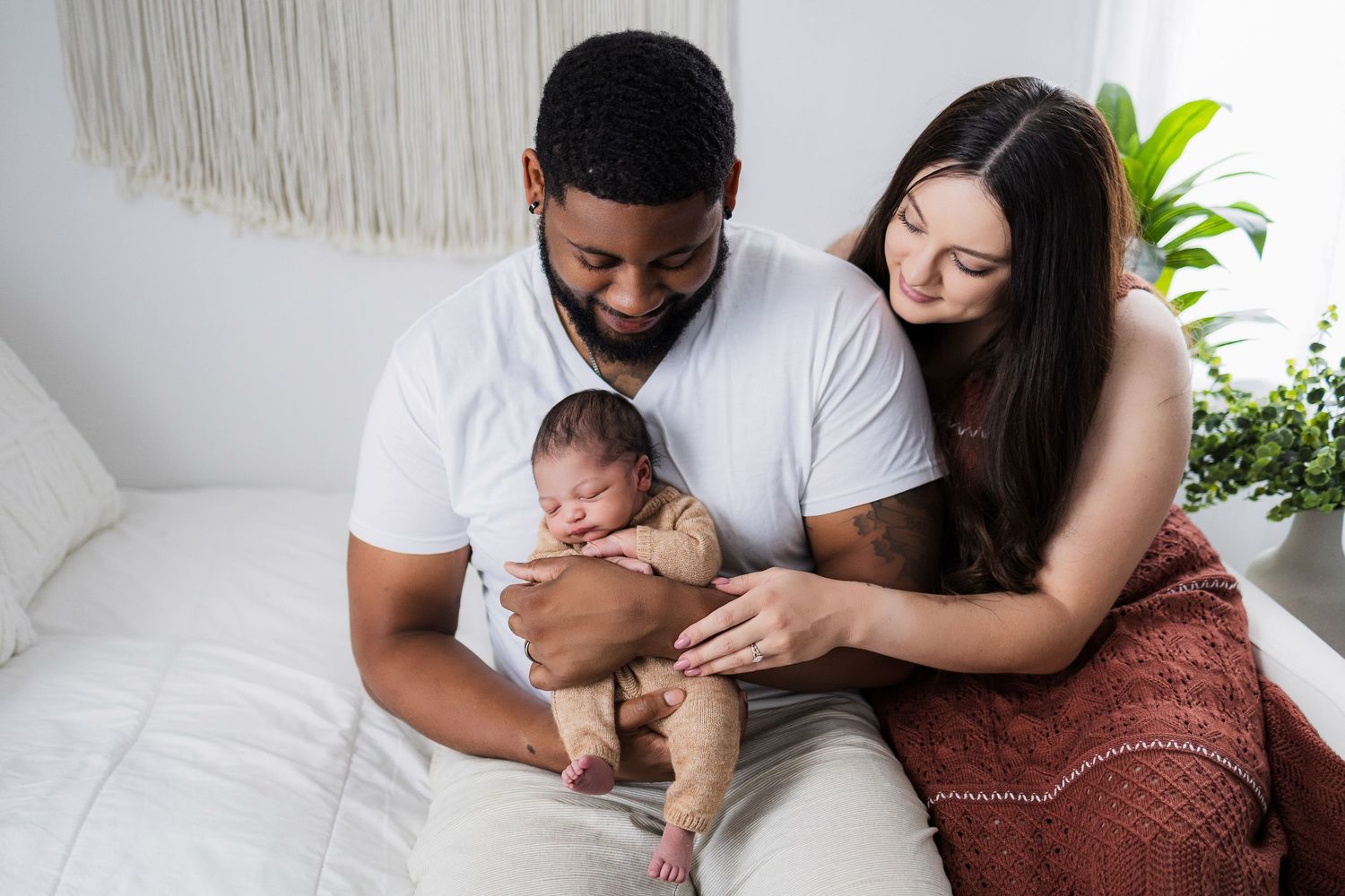 Parents sitting on white couch tenderly holding their newborn baby while surrounded by minimalist decor and plants.