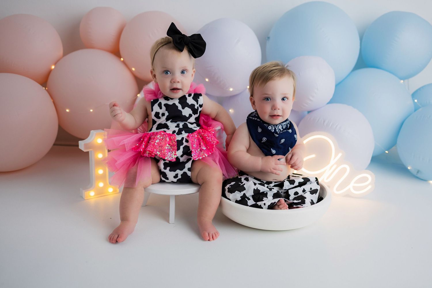 Two babies in matching black and white floral outfits sit against pastel balloon backdrop with illuminated number one.