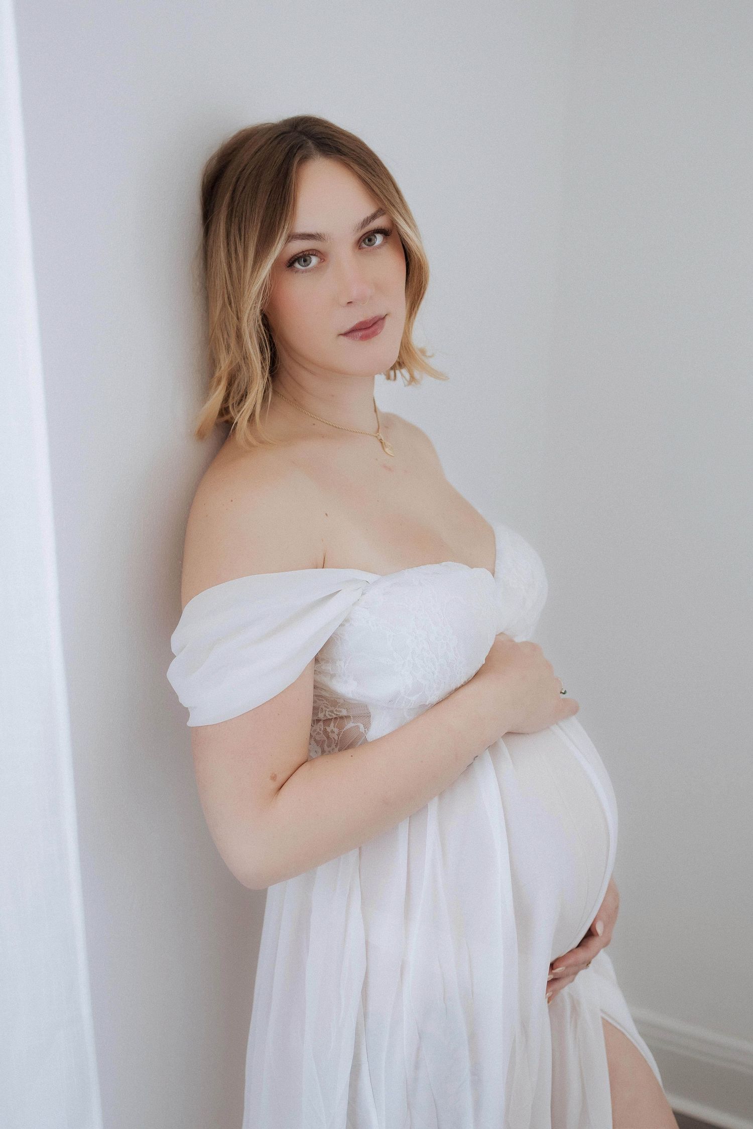 Maternity photo of woman in white off-shoulder dress against light wall.