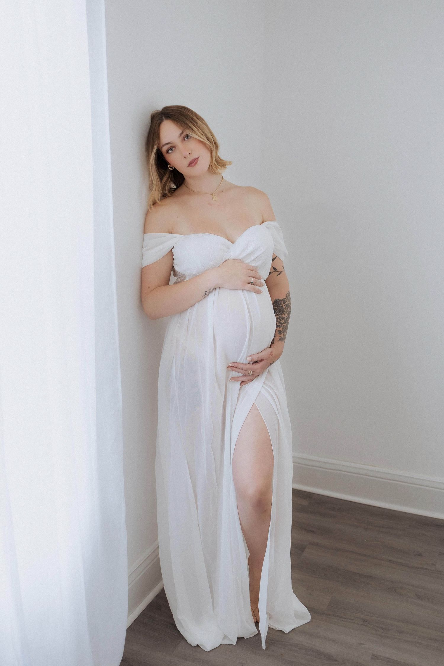 An elegant off-shoulder white maternity dress with thigh slit poses against light grey wall in a minimalist setting.