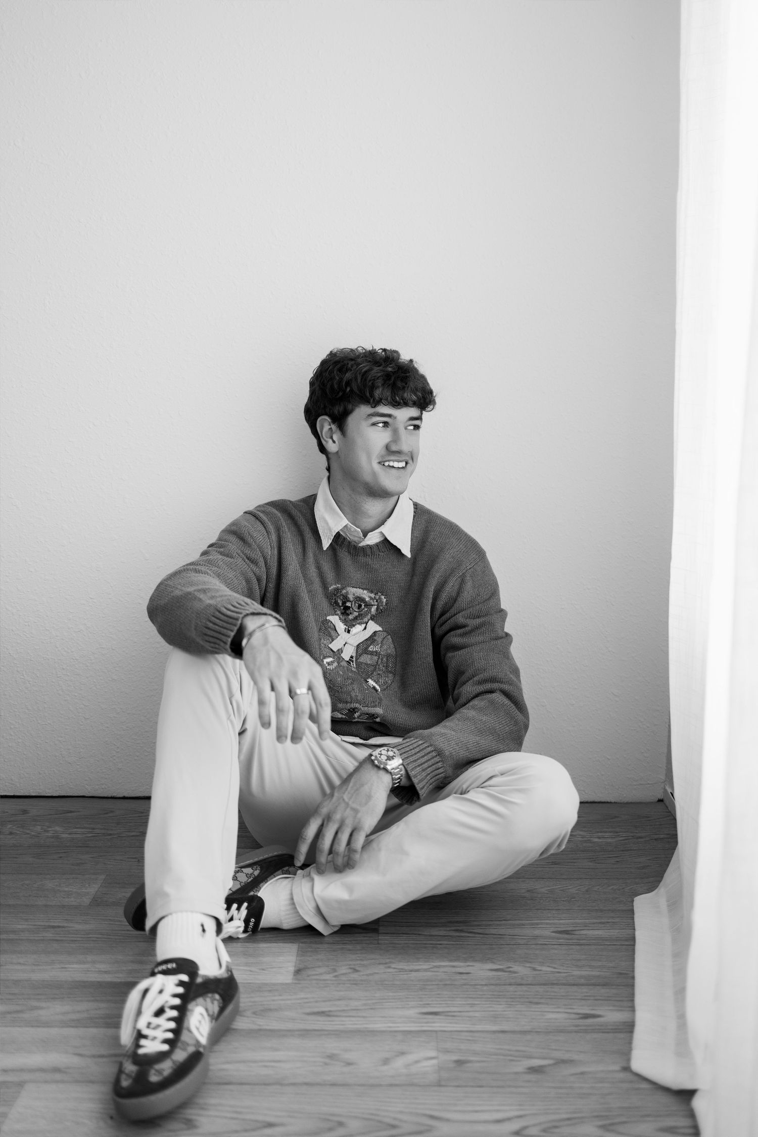 Black and white photo of a young man sitting cross-legged on a wooden floor, wearing a graphic sweater and sneakers.