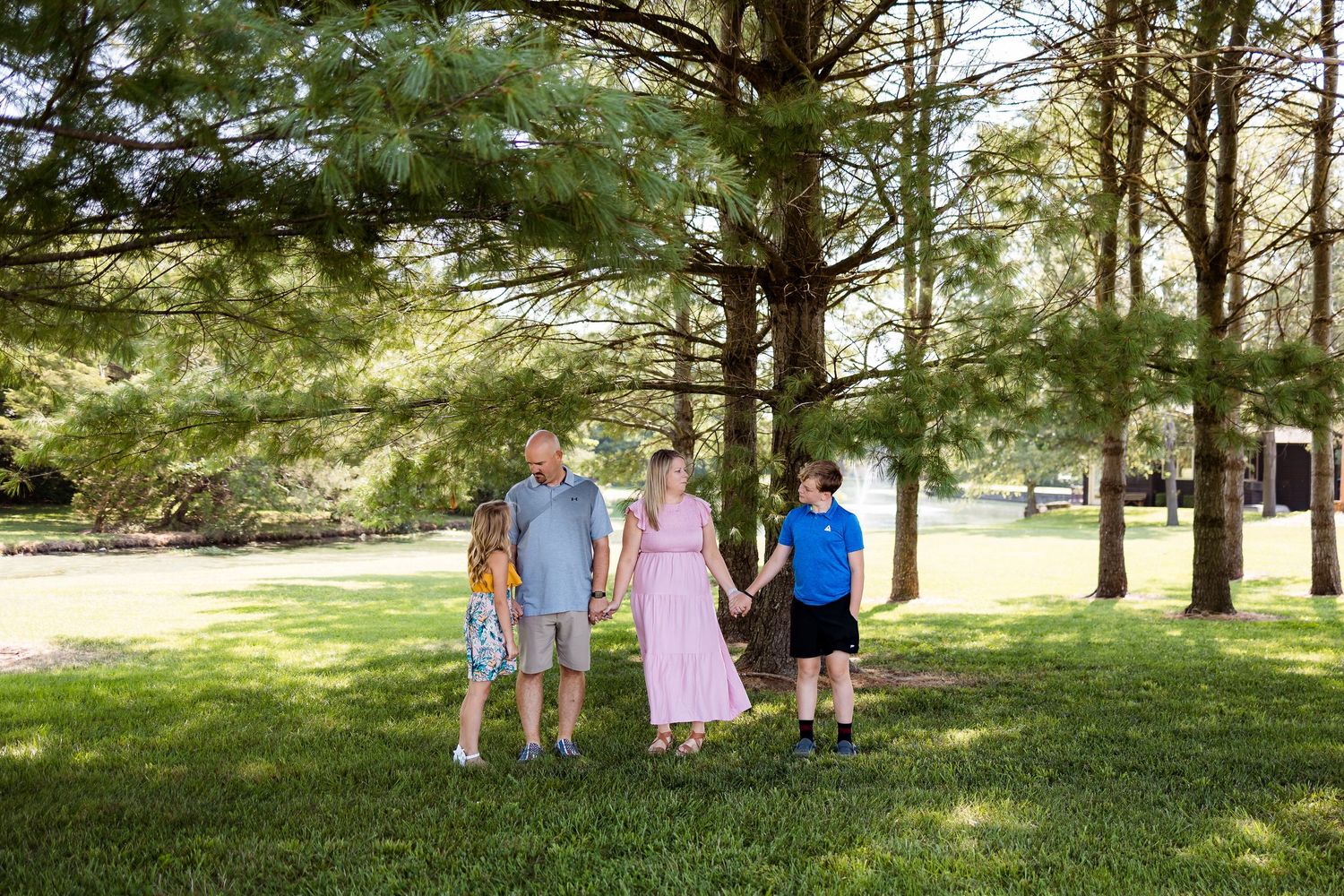 Family group stands together outdoors wearing casual summer attire among tall evergreen trees in a park setting.