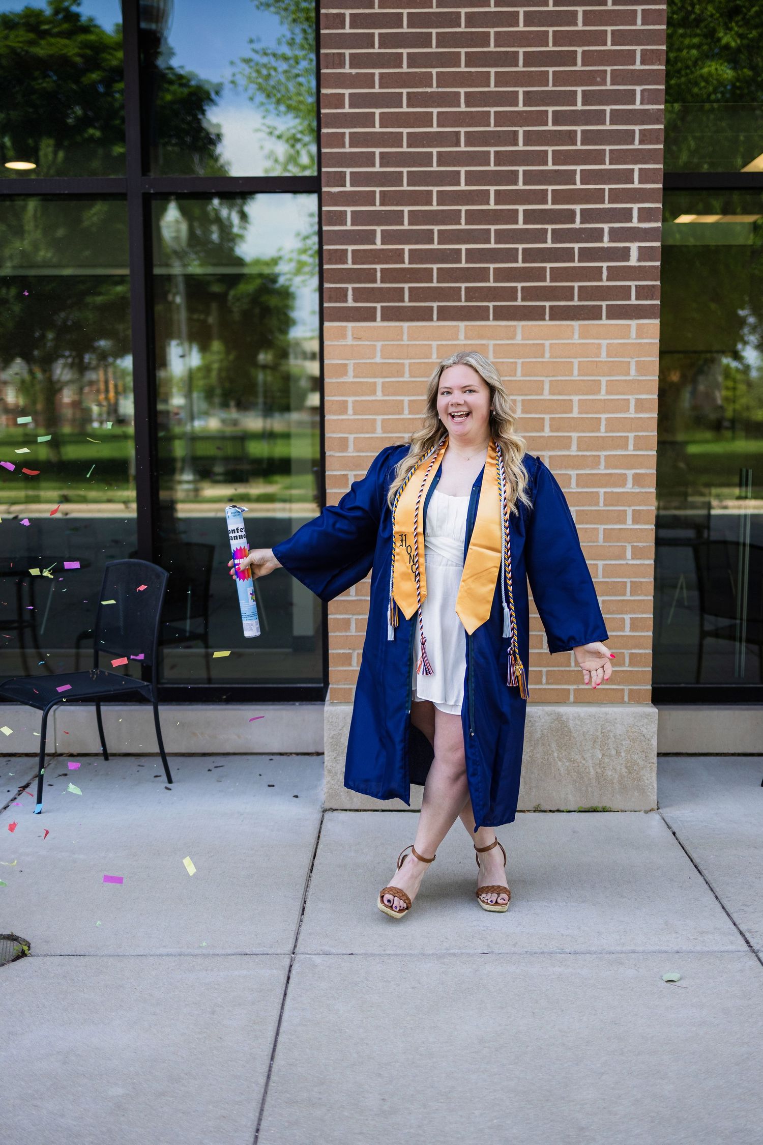 Graduate in blue cap and gown holding diploma against brick wall with reflective windows and outdoor seating.