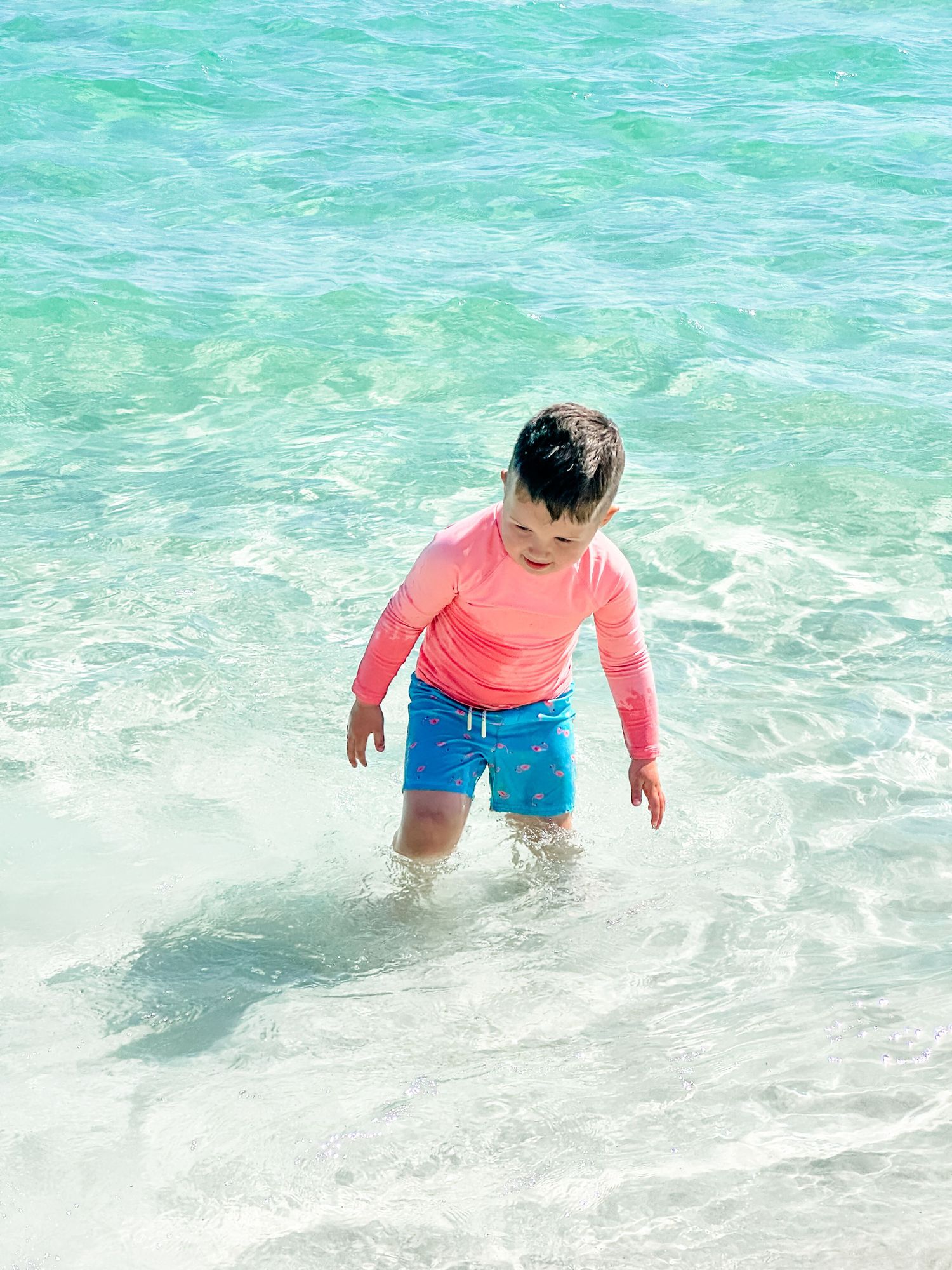 Child wading in crystal clear turquoise waters wearing pink and blue beachwear.