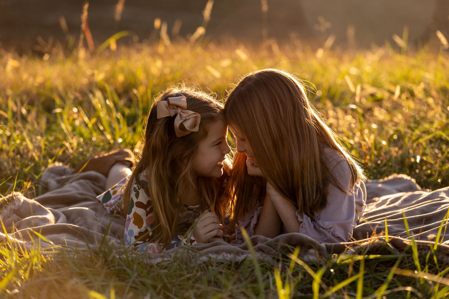 Sisters lying on blanket in golden sunlit grass during sunset.