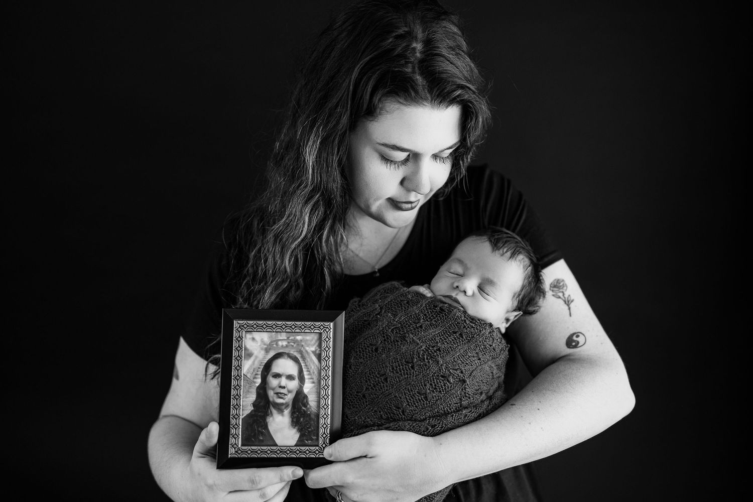 Black and white artistic portrait of a person holding both a sleeping baby and a framed photograph against dark background.