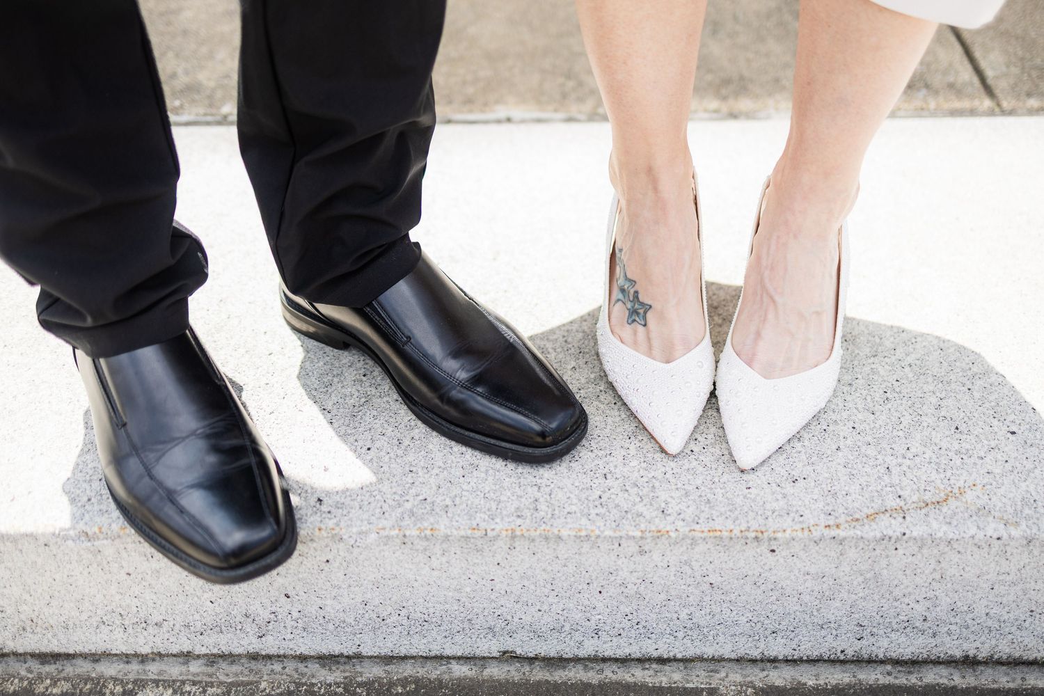 Wedding couple's feet showing black leather dress shoes and white bridal heels on concrete step.