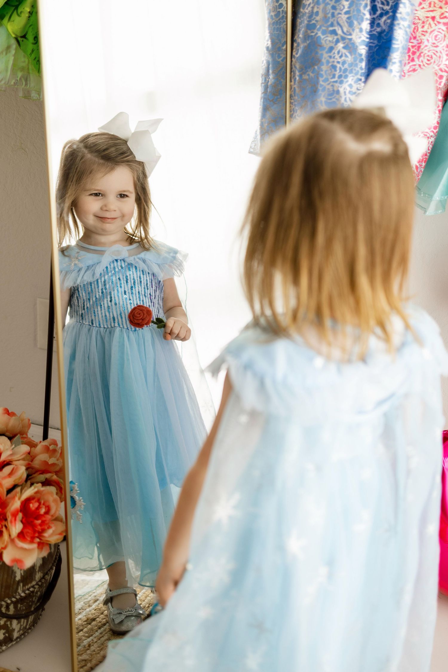 A child in a light blue dress and white bow looks at their reflection in a mirror with colorful decorations nearby.