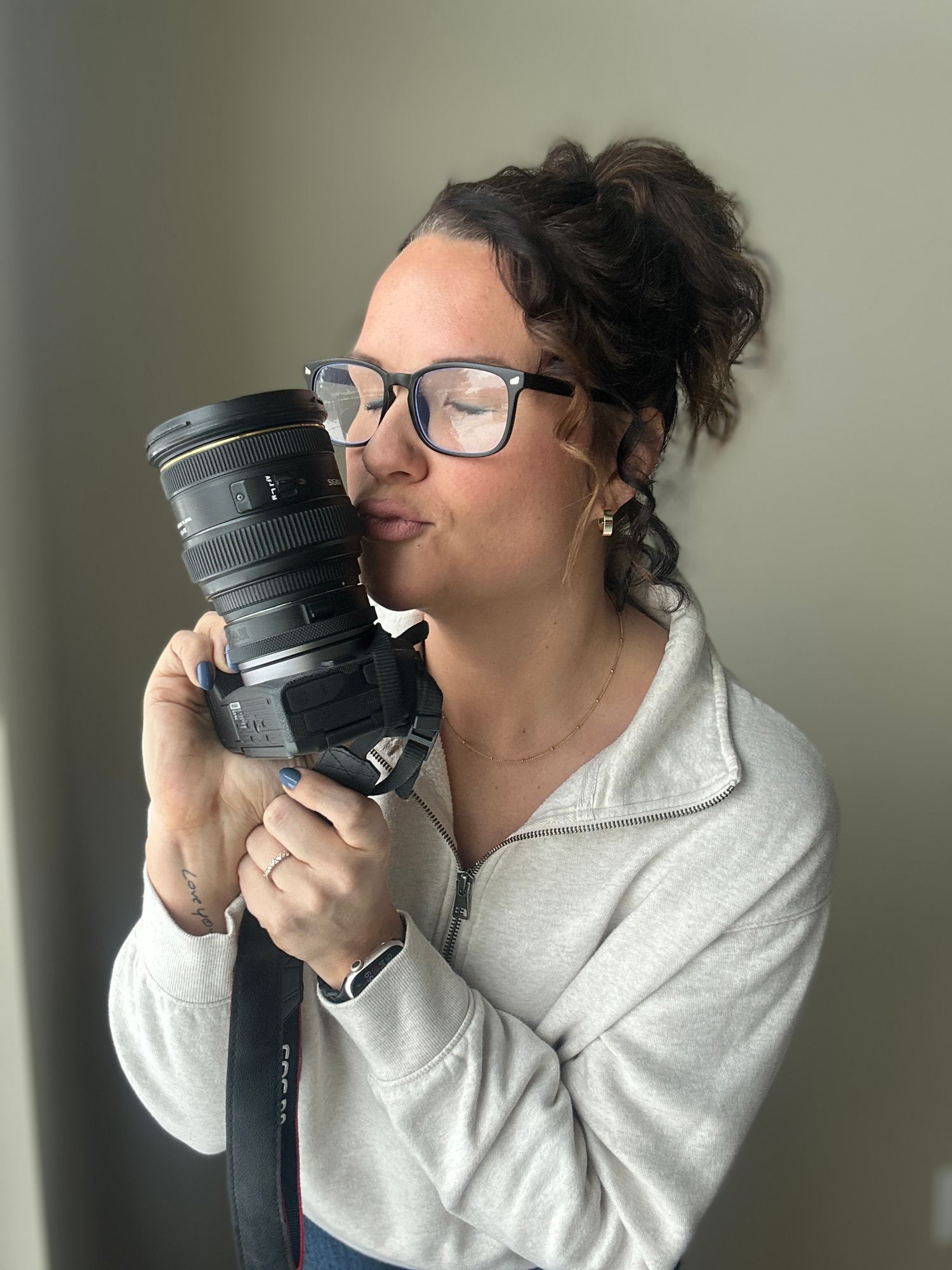 Person in glasses and white sweater affectionately hugging a professional camera lens against neutral background.