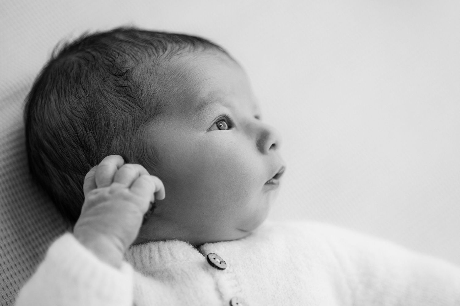 Black and white close up profile of a newborn baby in white swaddling looking to the side against light background.