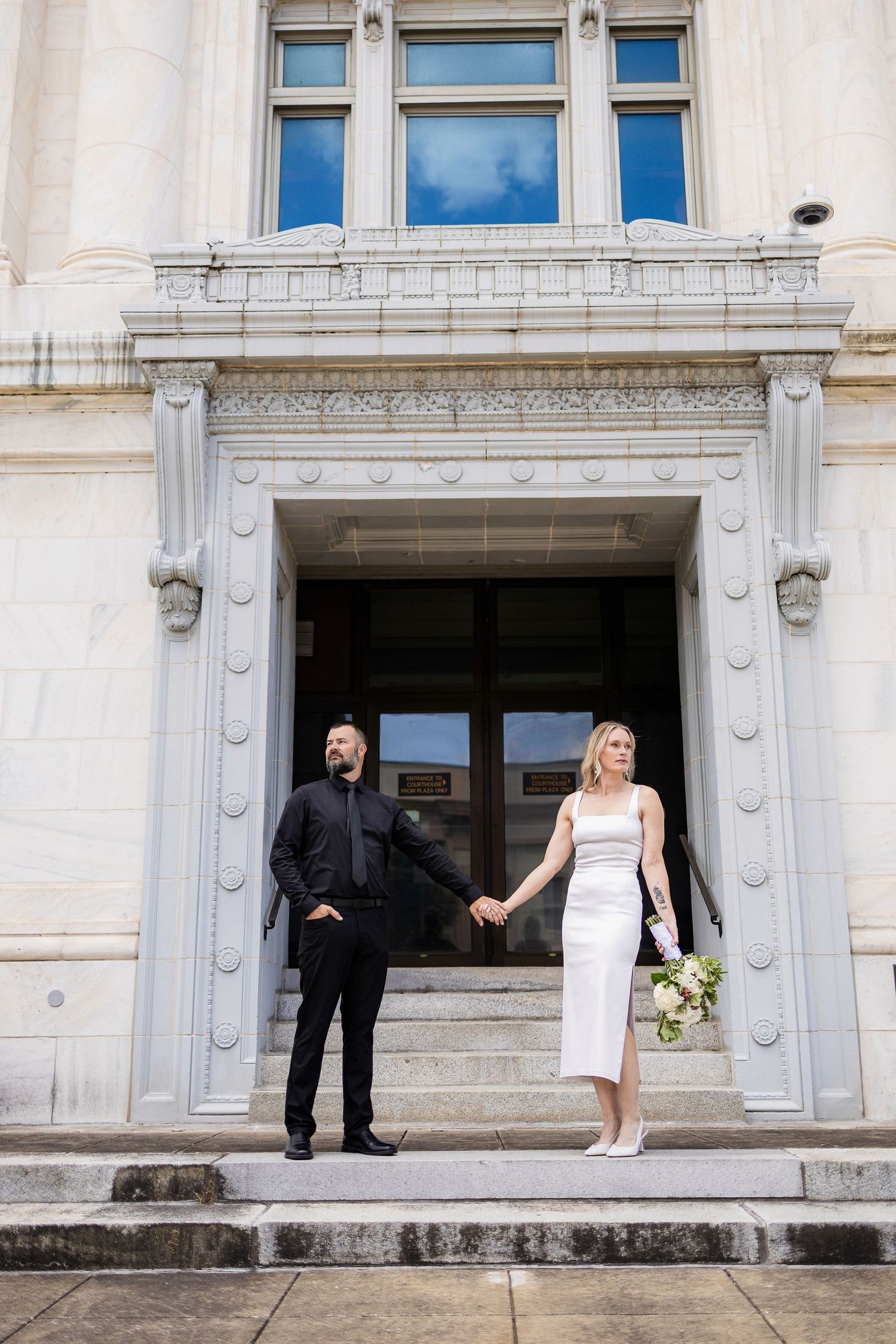 A couple stands on stone steps in front of an ornate white building entrance with classical architectural details.