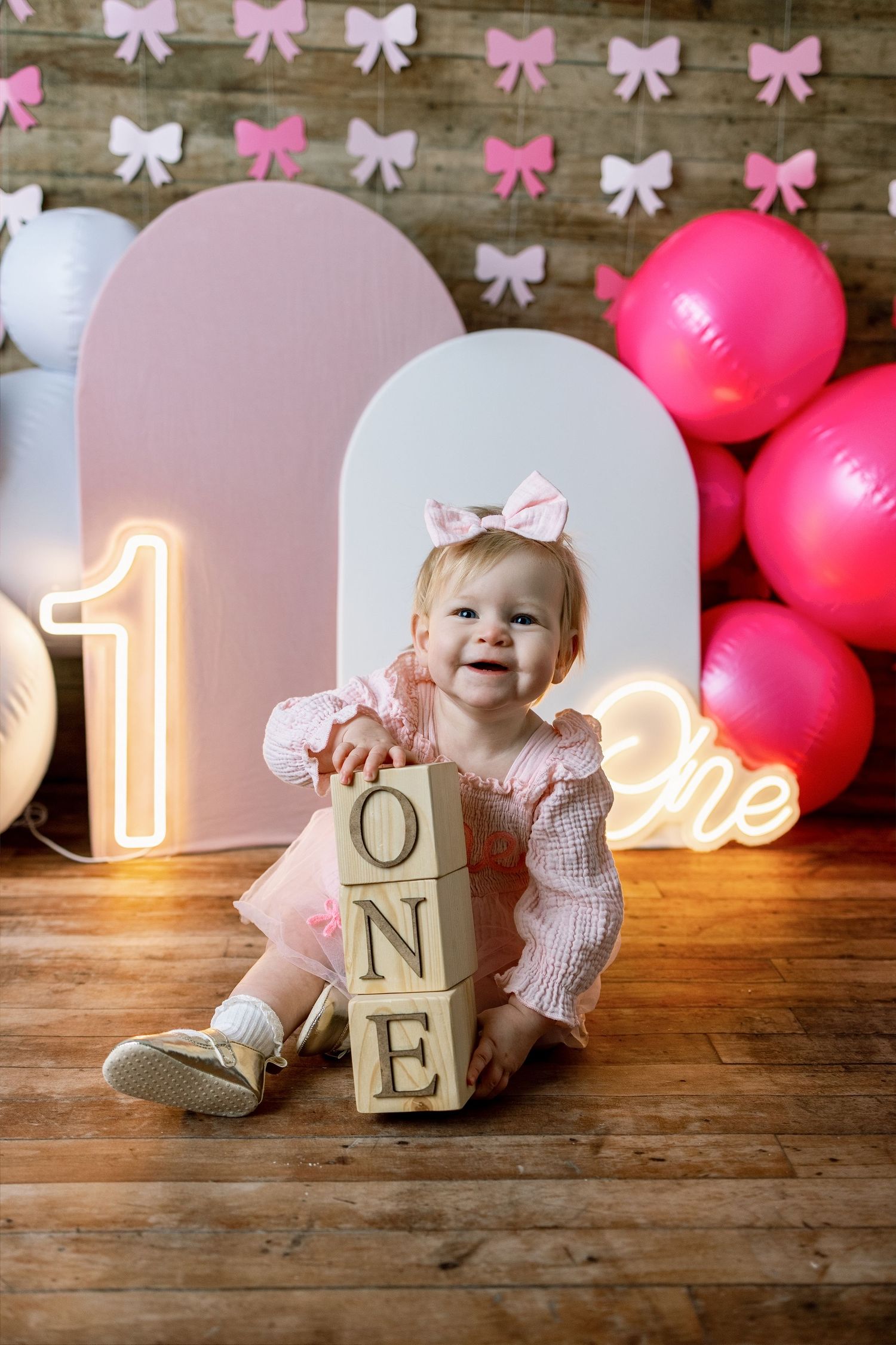 First birthday party setup with pink and white bows, wooden blocks, and glowing number one on rustic wooden floor.