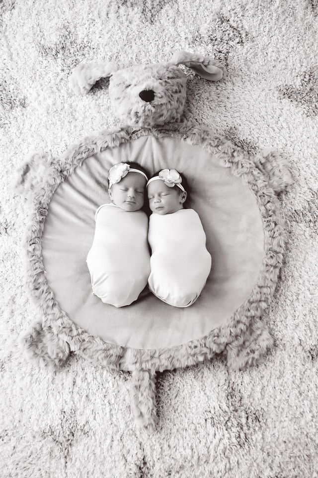 Two swaddled newborn babies lying together on a round cushion with white carpet background in black and white photography.
