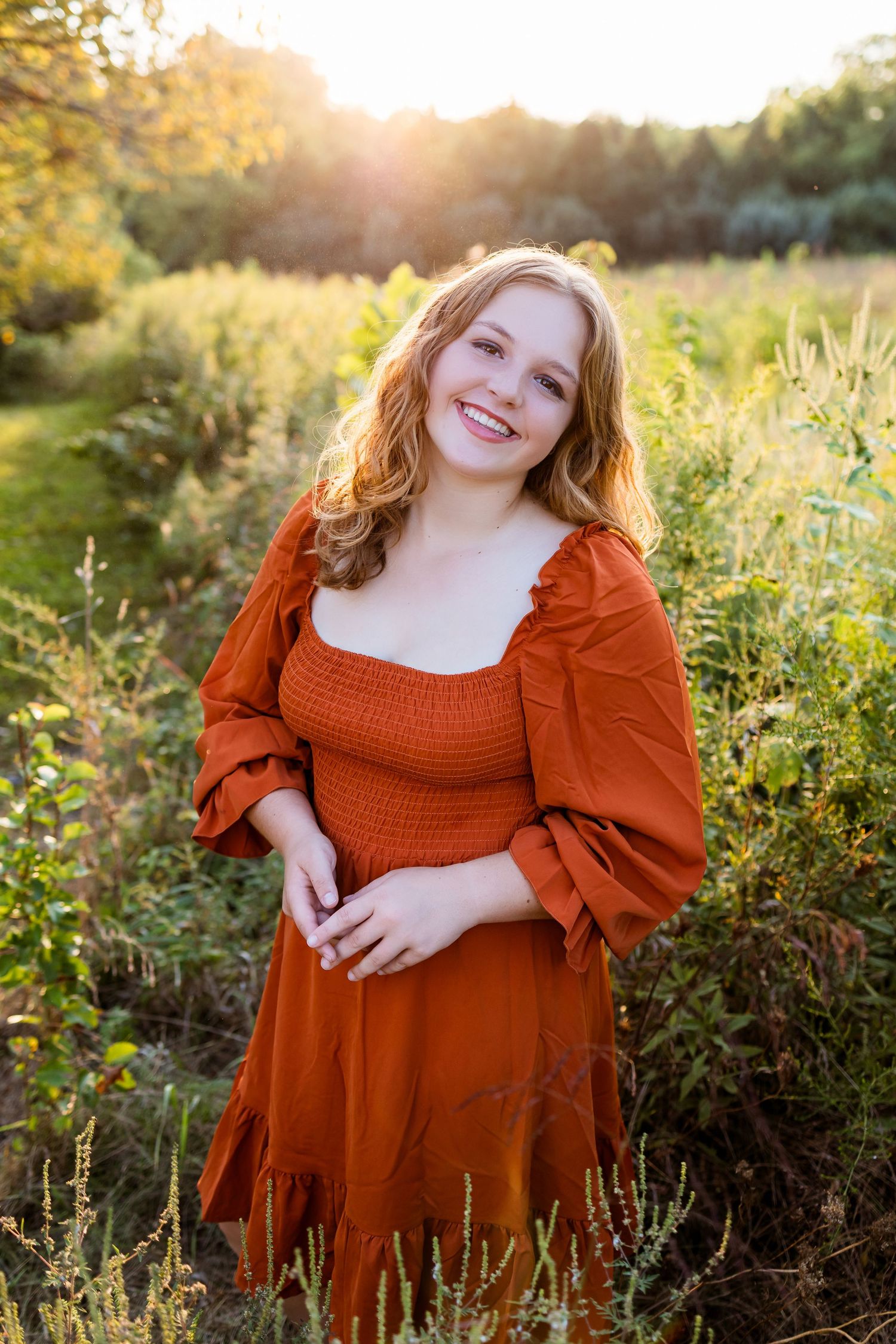 A person in a rust-colored dress poses in a golden sunlit field during sunset with wildflowers and tall grass.