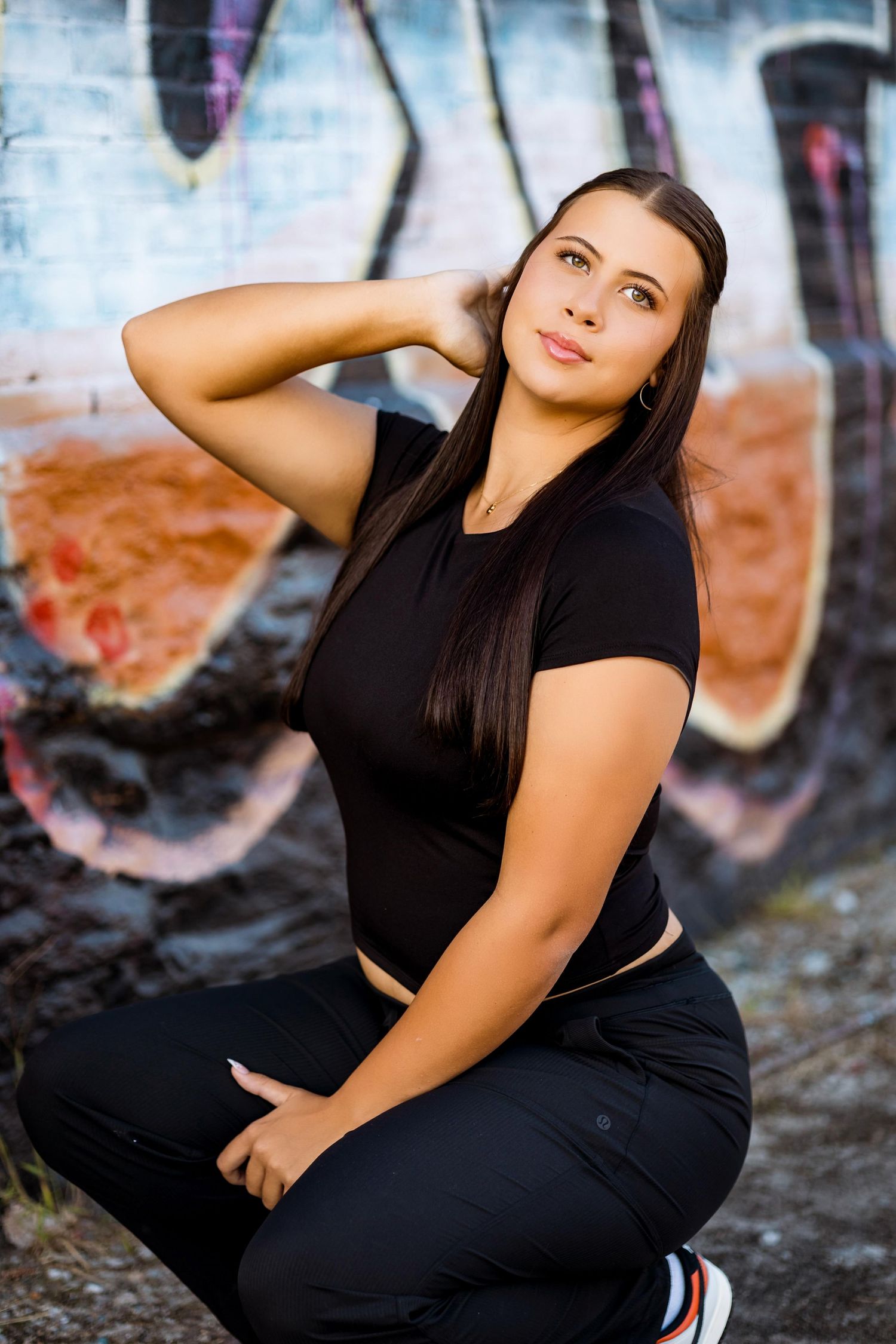 A person in all black athletic wear poses against a graffiti wall background.