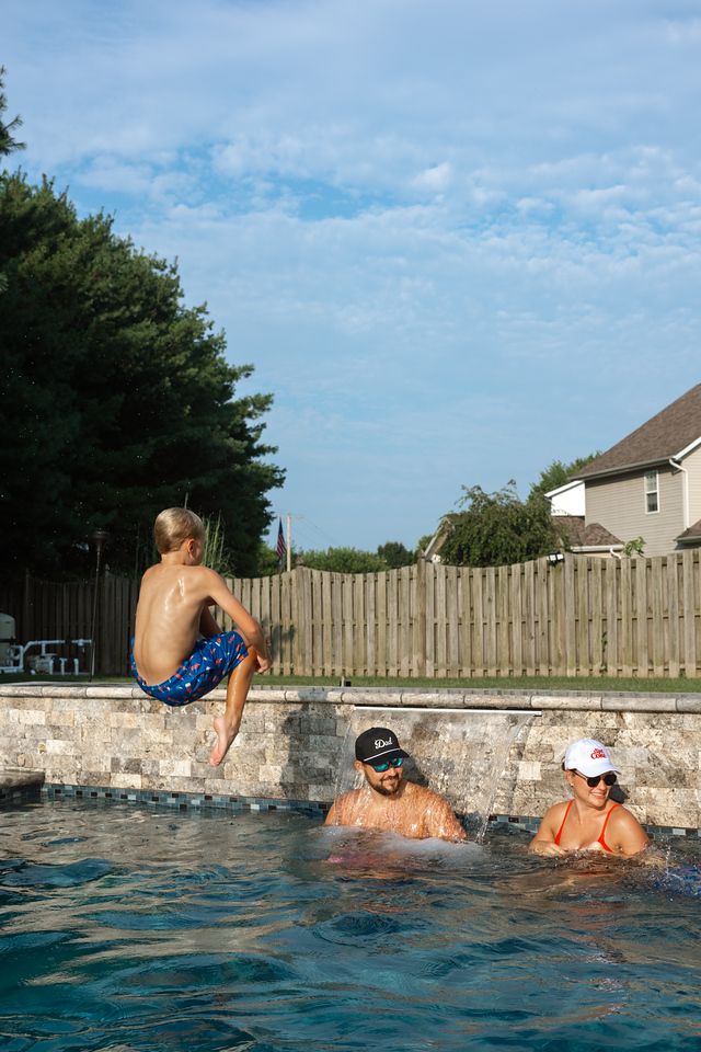Boy jumping into backyard pool while two adults in caps watch from the water on a sunny day.