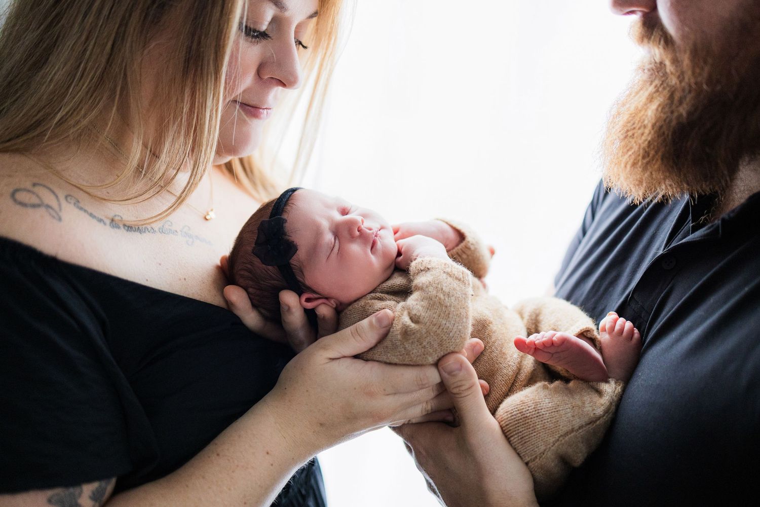 New parents tenderly cradle their sleeping newborn baby wrapped in beige blanket near bright window.