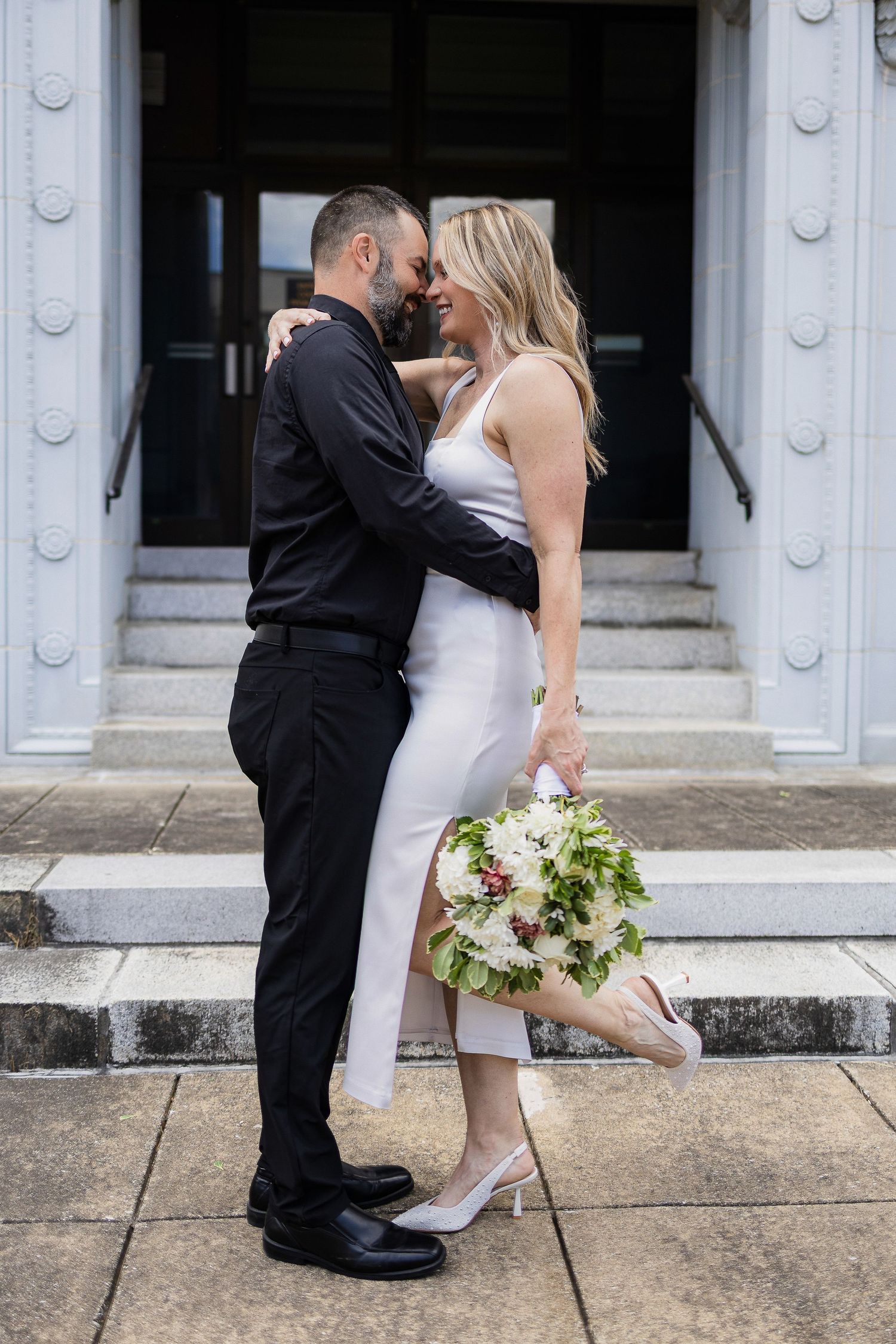 Romantic couple embraces on steps with white bouquet, bride in tea length dress and groom in black attire.