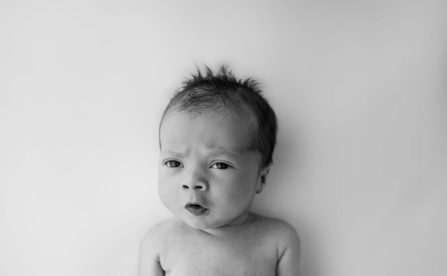 Black and white portrait of a newborn baby with dark hair lying on a white background, looking up with wide eyes.