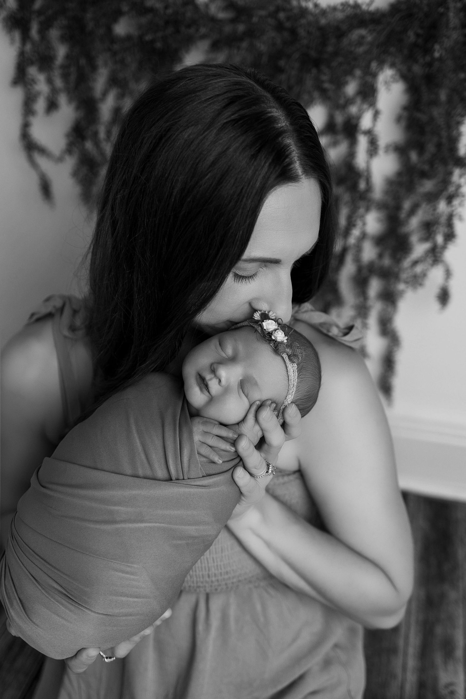 A tender black and white portrait of a mother lovingly embracing and kissing a sleeping newborn swaddled in fabric.