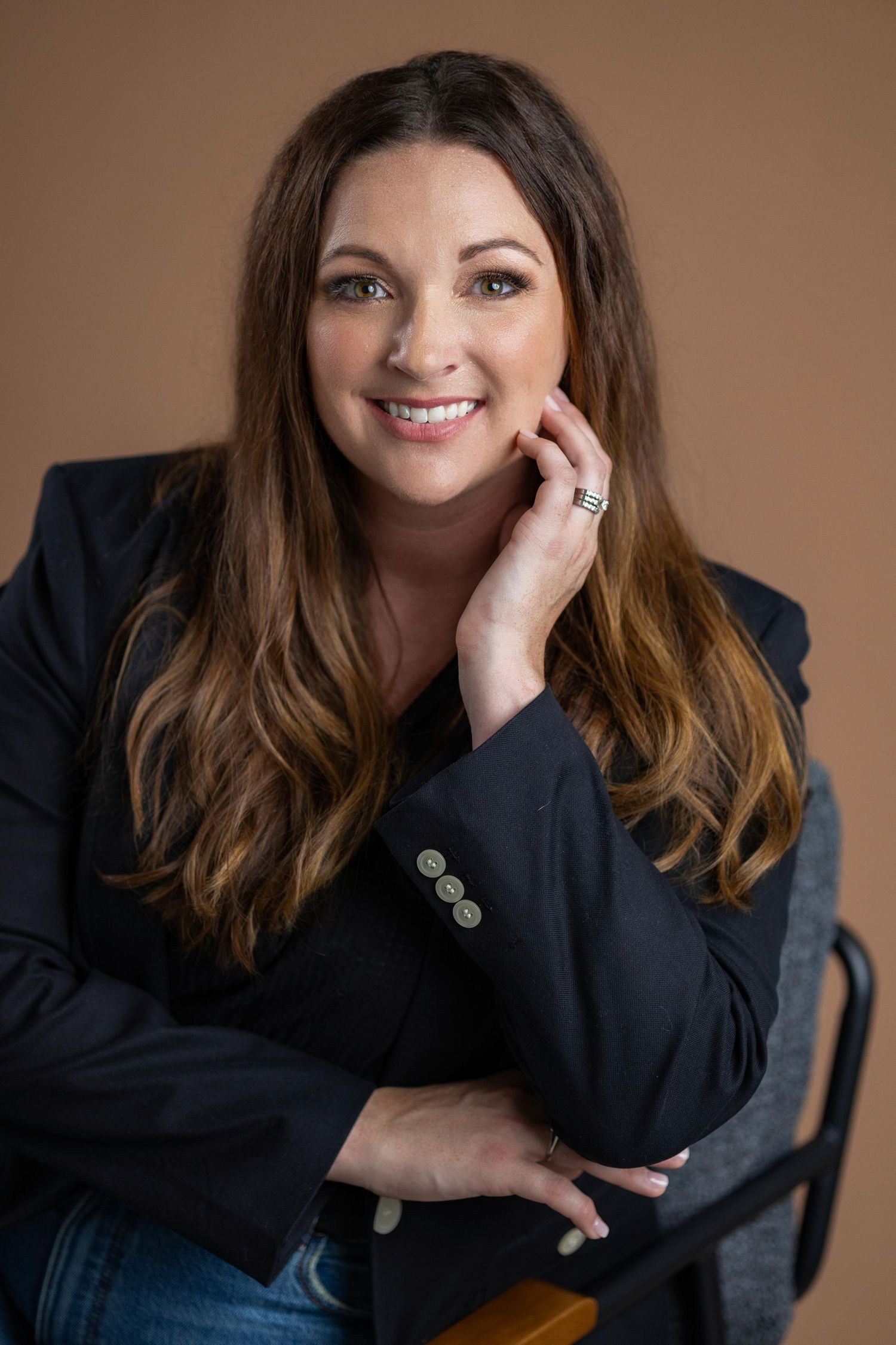 Person in dark blazer poses professionally against neutral brown background in studio setting.