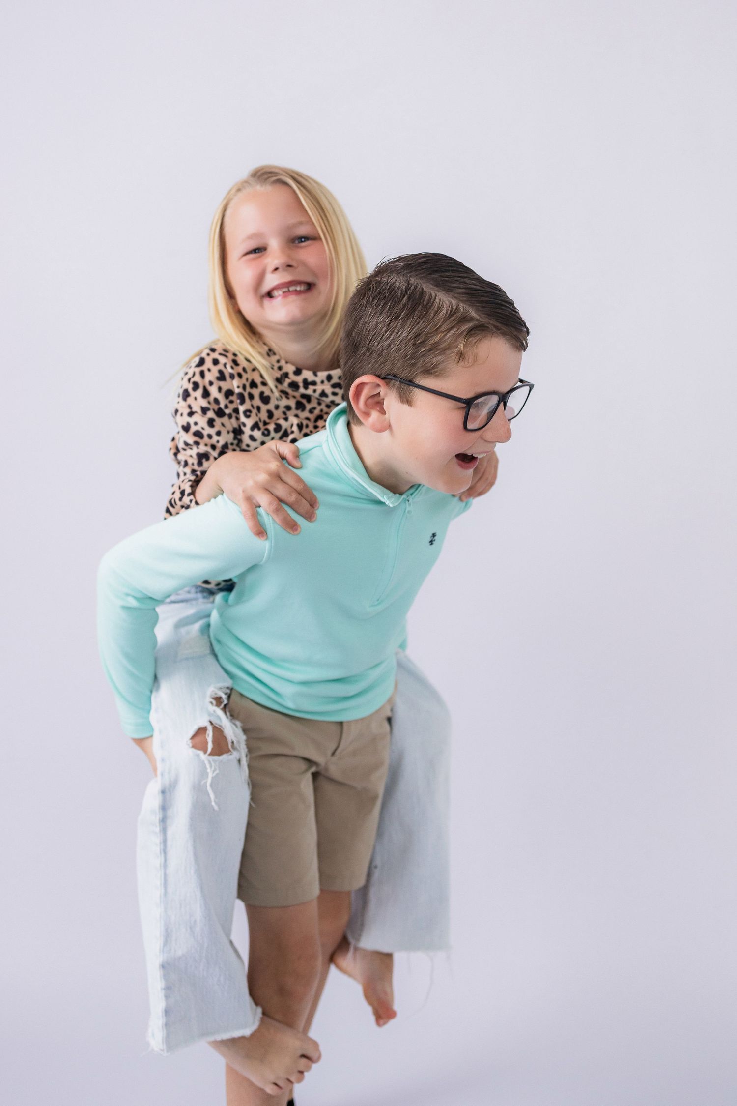 Two young children play and laugh together against a white background, with one giving the other a piggyback ride.