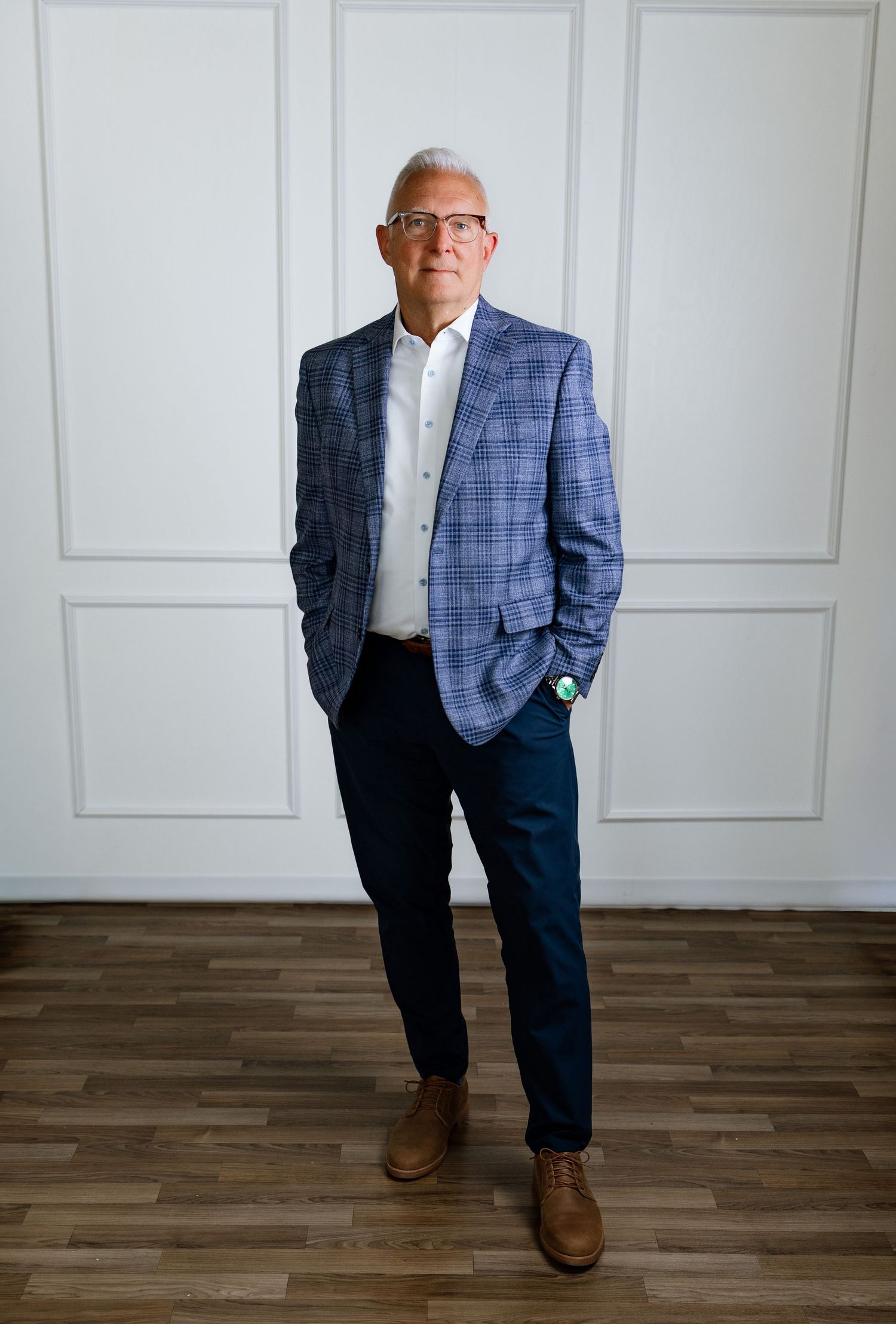 Professional man in blue plaid blazer and navy trousers standing confidently in a modern white interior.