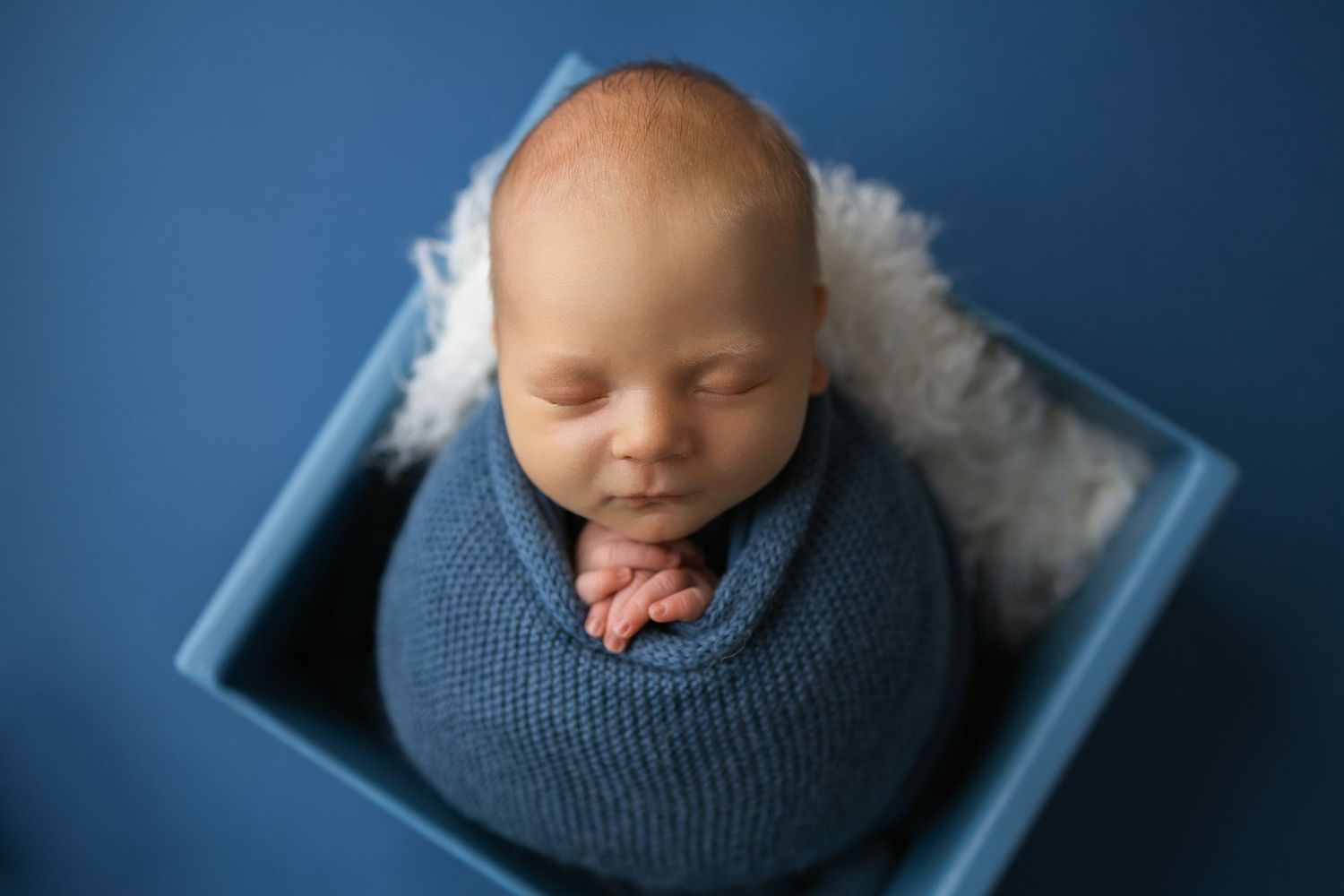 Newborn baby wrapped in blue knit blanket sleeps peacefully against dark blue background in professional portrait.