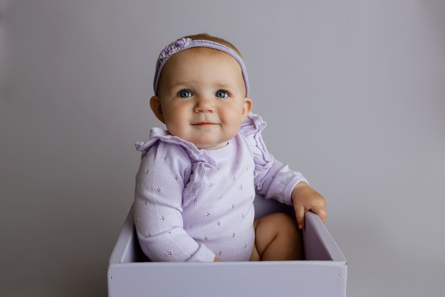 Smiling baby girl in lavender onesie and headband sitting in a white box against a gray background.