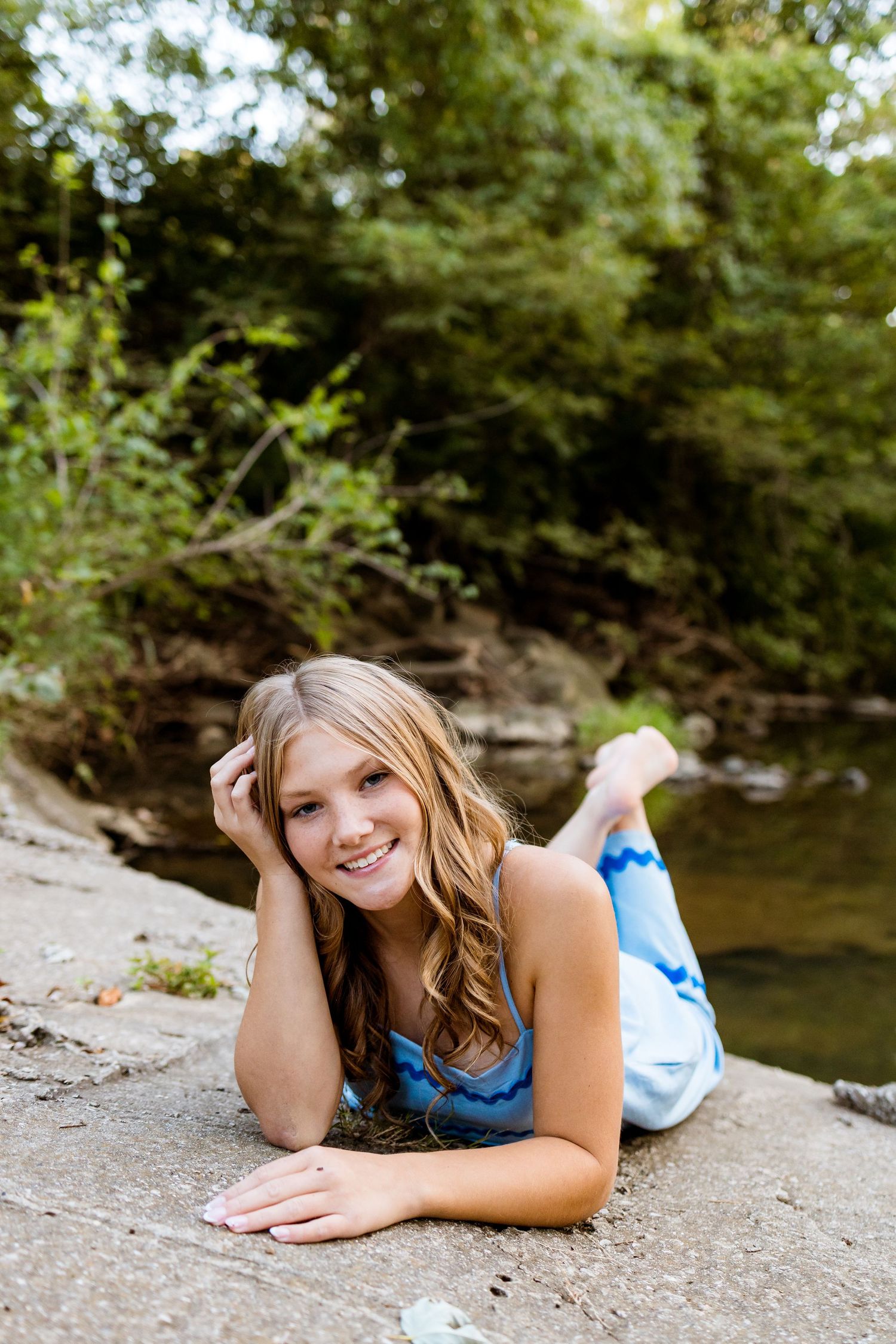 A person in a blue top lying on a rocky riverbank surrounded by lush greenery on a sunny day.