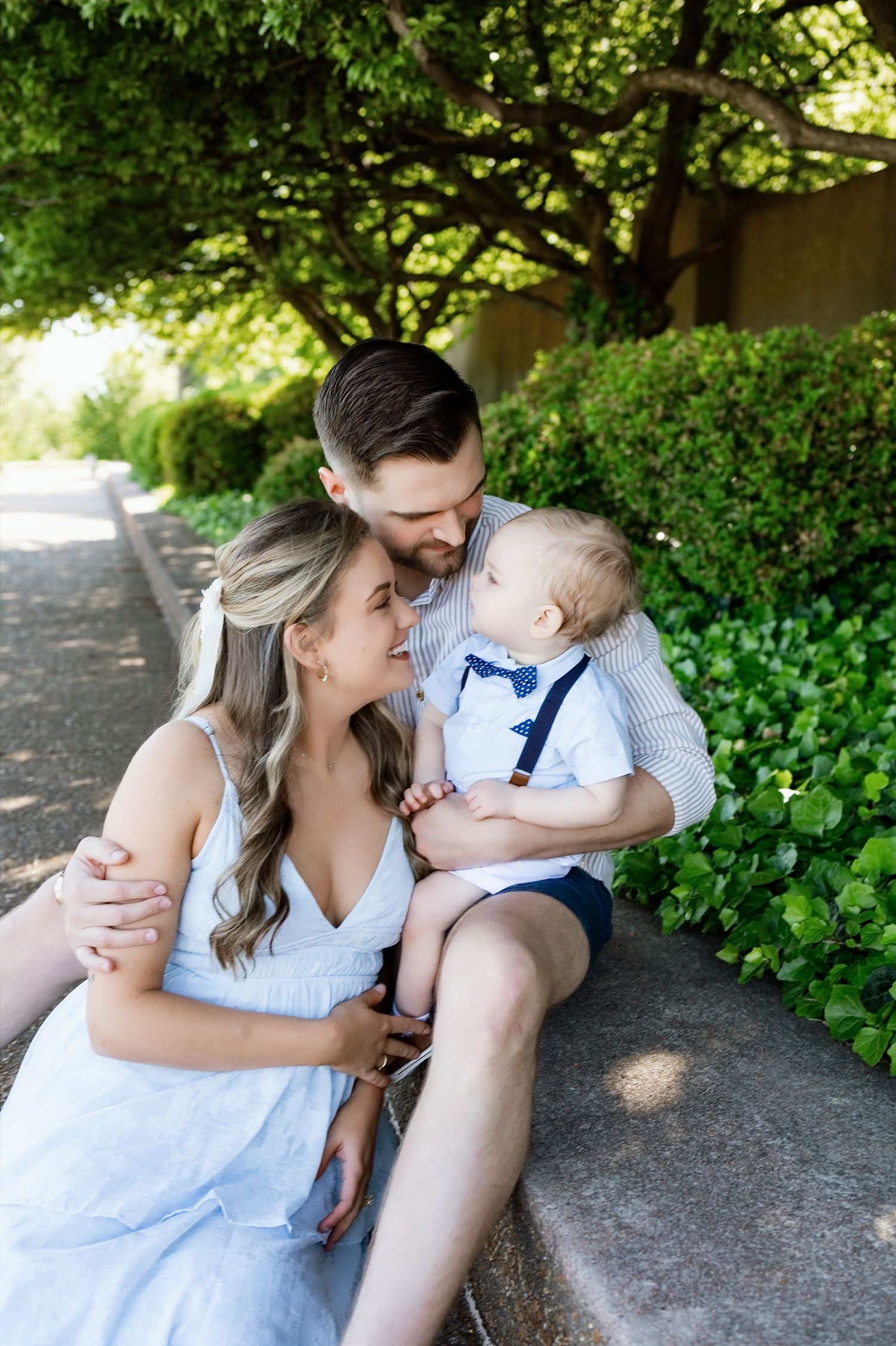Loving couple sharing tender moment with infant son in blue bow tie and suspenders on outdoor stone steps.