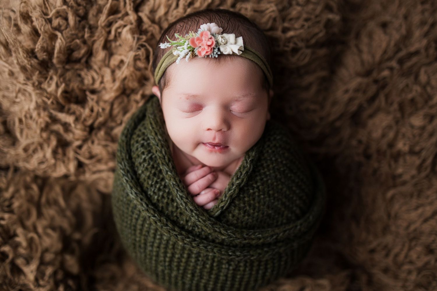Sleeping newborn wrapped in cozy green knit blanket with delicate pink floral headband in artistic portrait series.