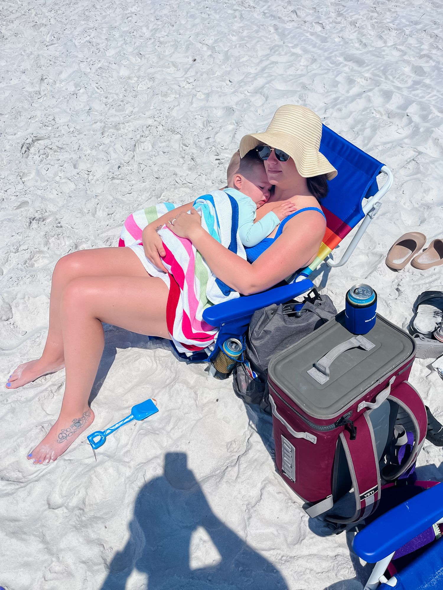 Person relaxing in a beach chair with sunhat and striped dress next to a cooler on white sand.