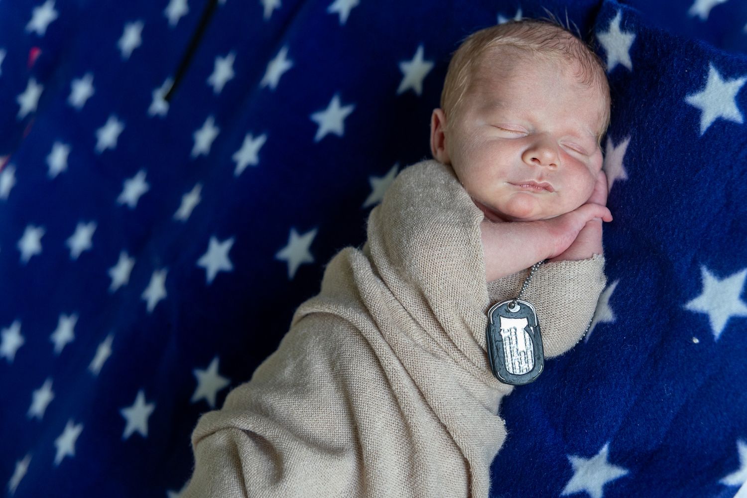 Sleeping newborn wrapped in beige swaddle blanket on blue star-patterned background with military dog tags.