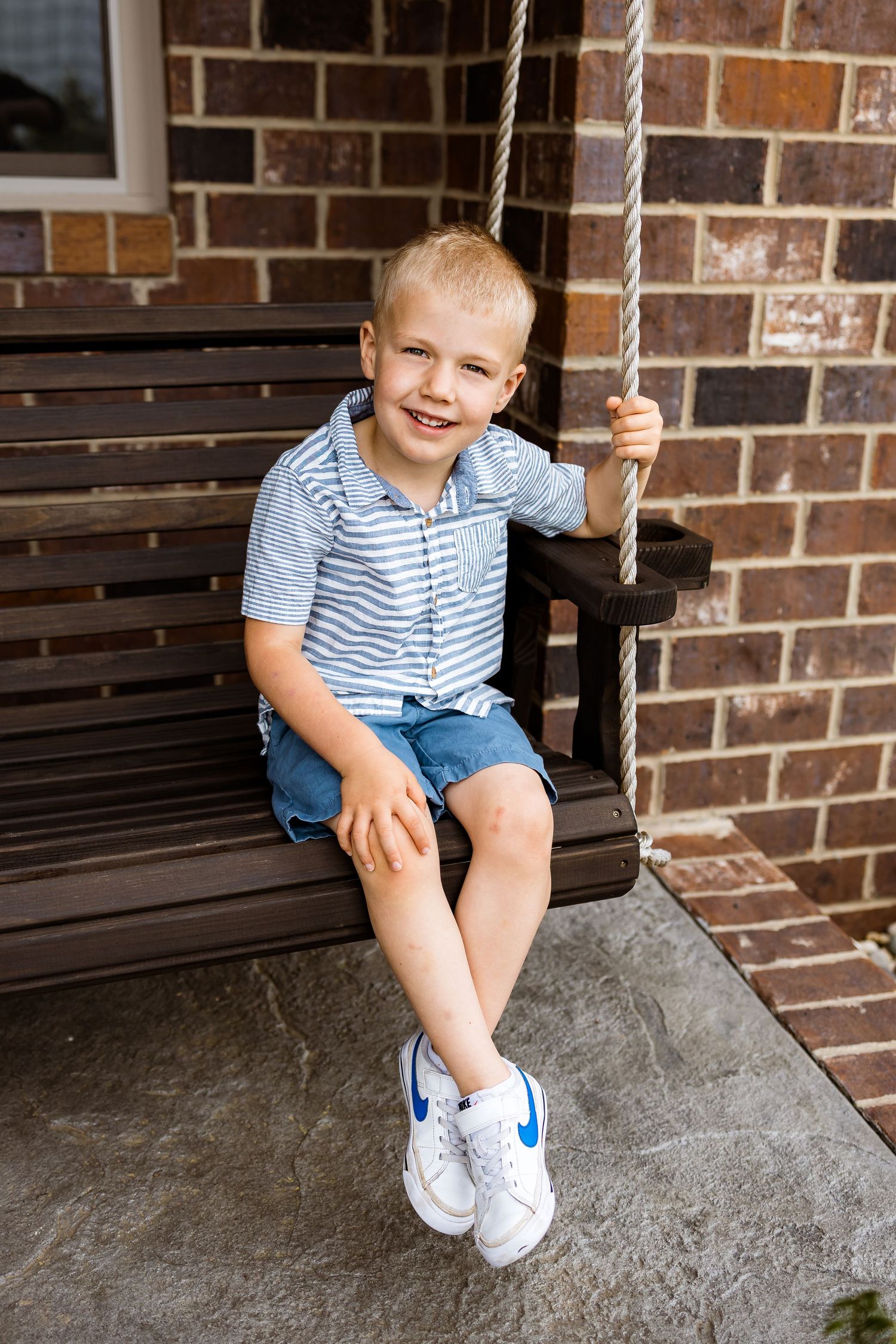 Young boy in striped shirt sitting on porch swing by brick wall.