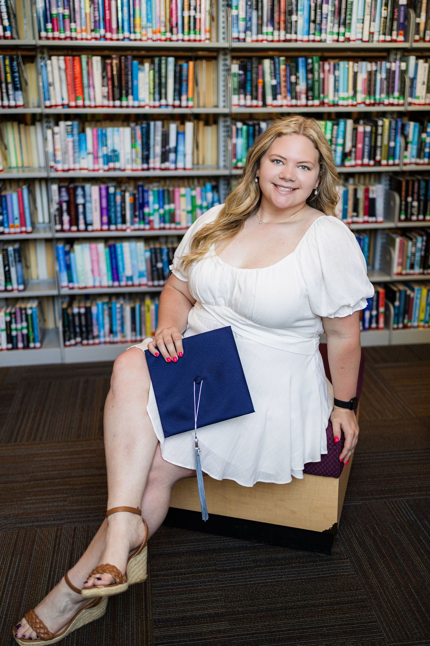 Graduate in white dress poses in university library with blue diploma holder against backdrop of bookshelves.