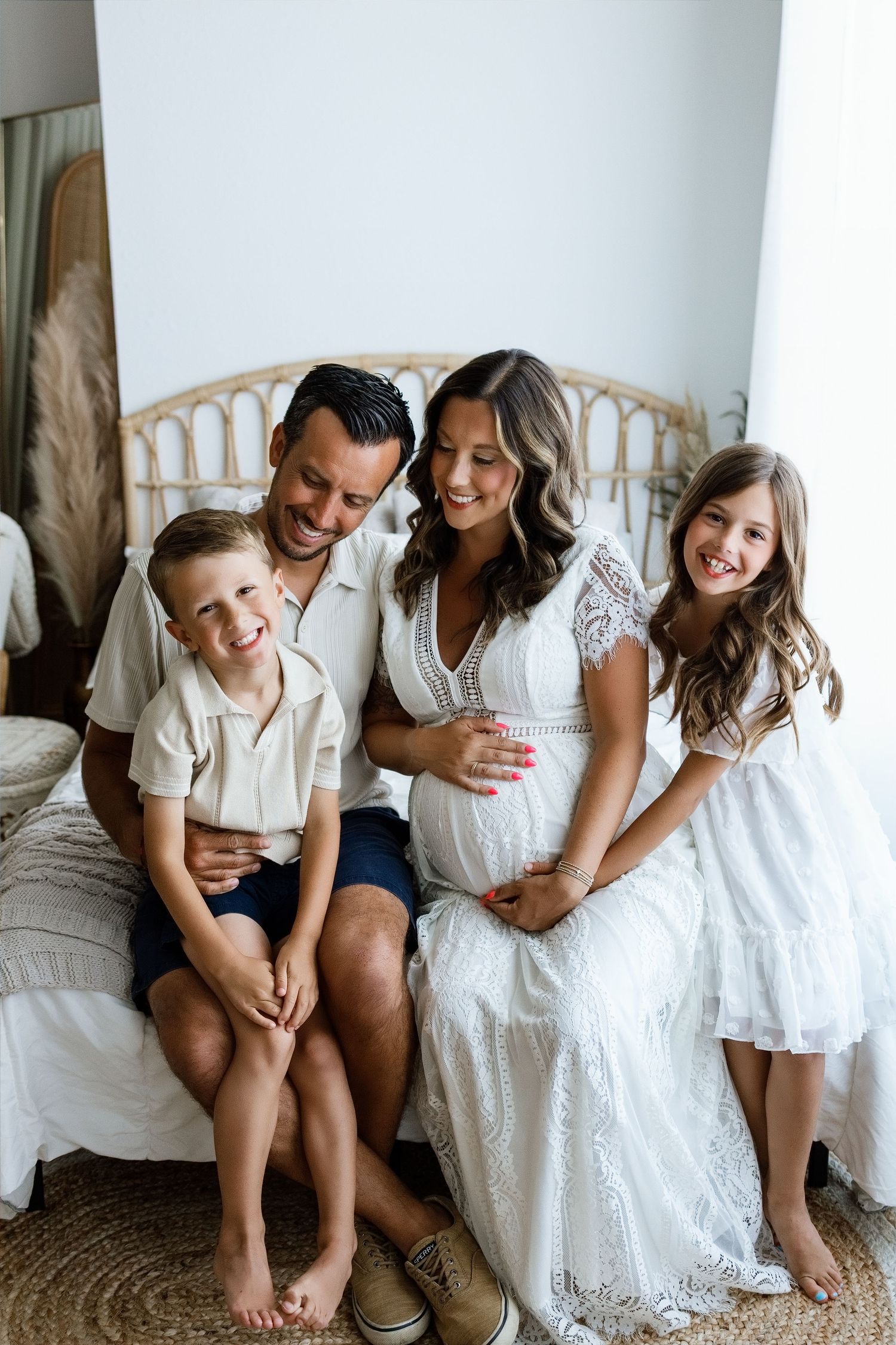 Happy family maternity photo with pregnant mother in white lace dress, father, son, and daughter sitting together on bed.