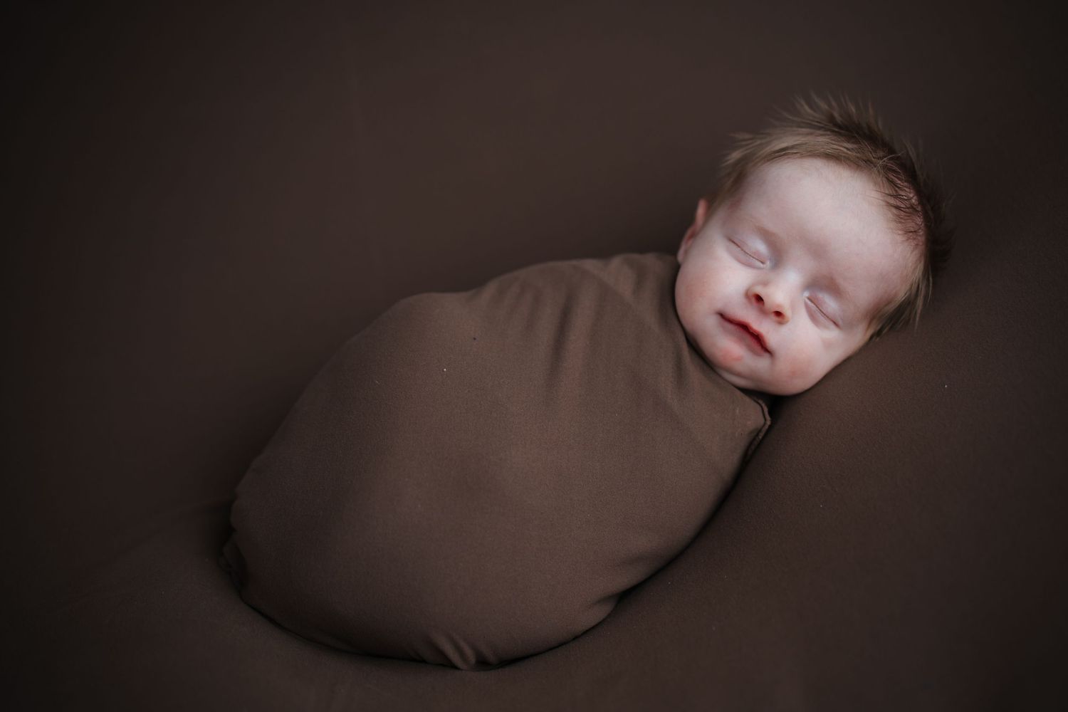 Peaceful sleeping newborn baby wrapped in brown swaddle against dark background.