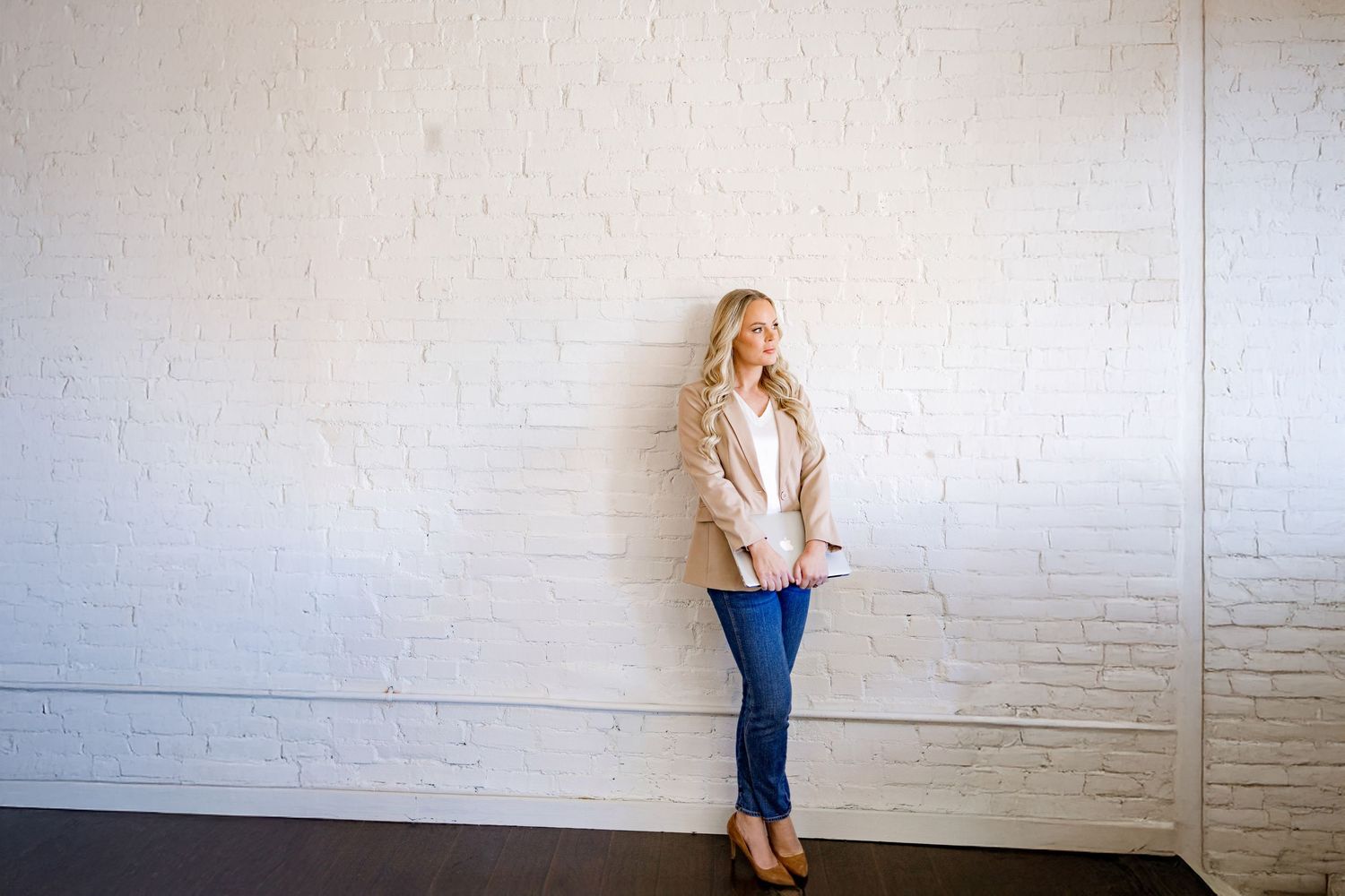 Woman in beige sweater and blue jeans stands casually against white brick wall with hands in pockets.