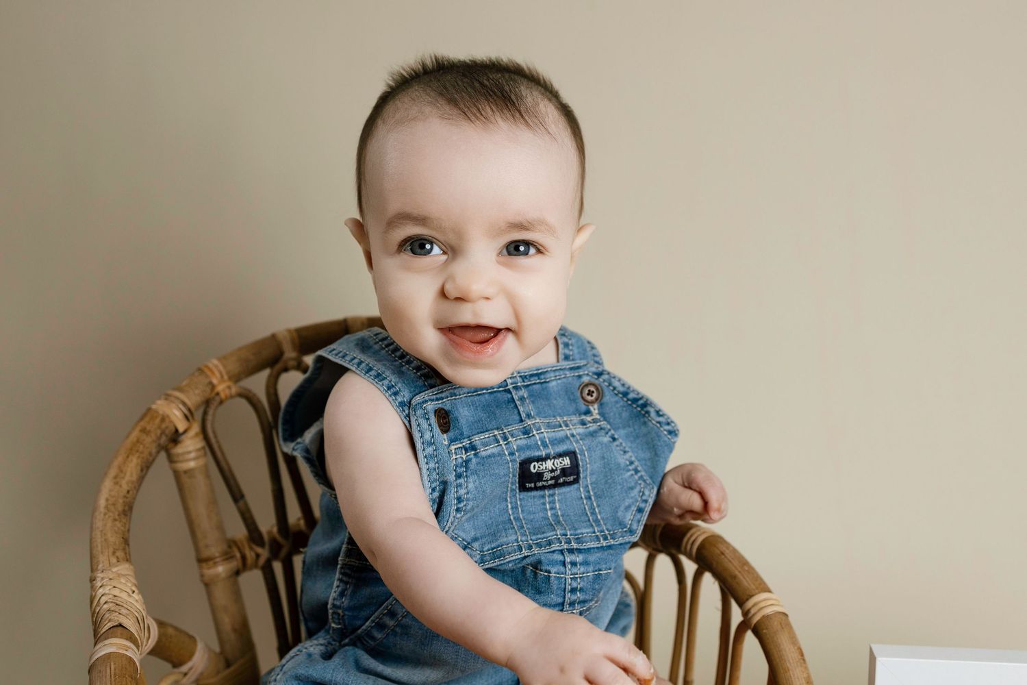 A happy baby wearing denim overalls sits in a wicker chair against a beige wall.