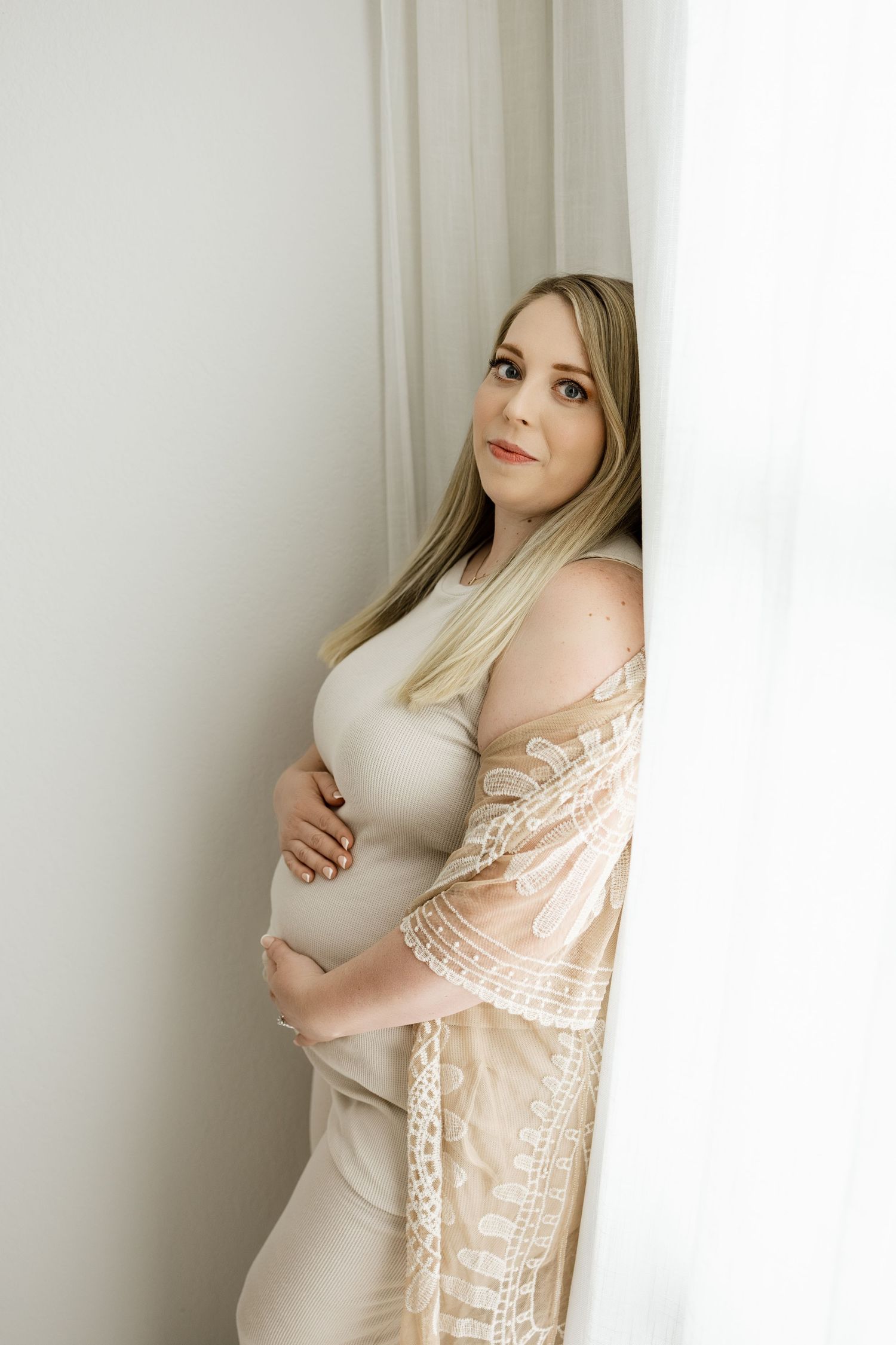 Ethereal maternity portraits in soft peach lace robe against white wall with natural window light.