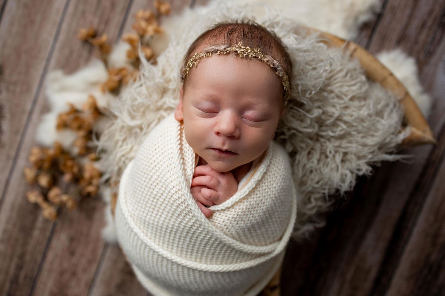Newborn baby wrapped in white knit blanket sleeping peacefully on fluffy rug with delicate gold headband.